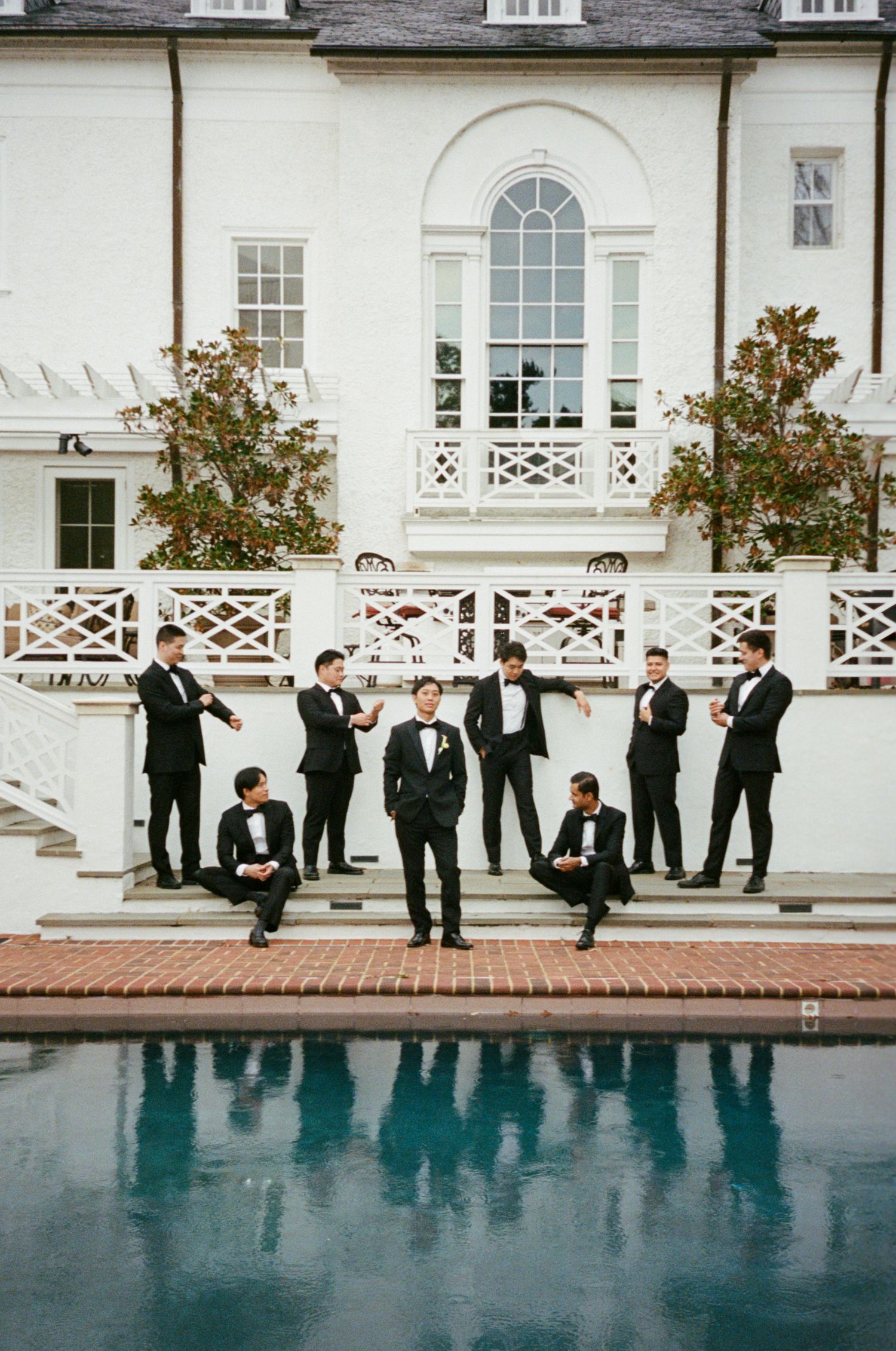 Wedding party in formal black tuxedos stands along an elegant poolside terrace at a white mansion.