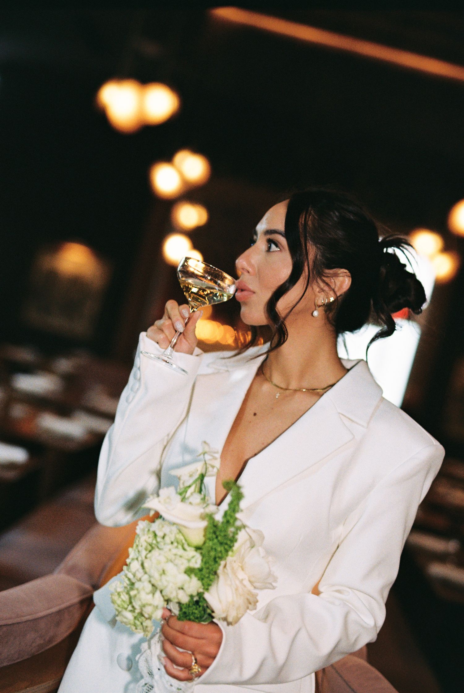 Bride in white blazer sipping champagne while holding white floral bouquet in elegant restaurant setting.