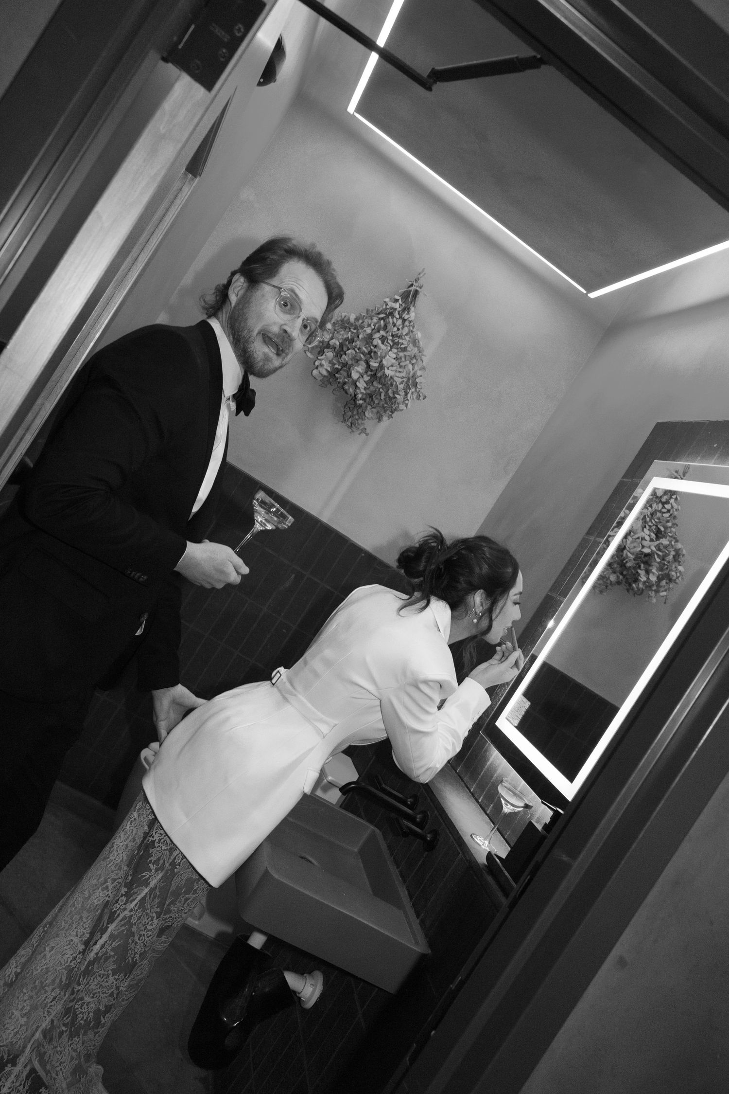 Black and white photo of a man in a tuxedo and woman in white dress at a bathroom vanity mirror.