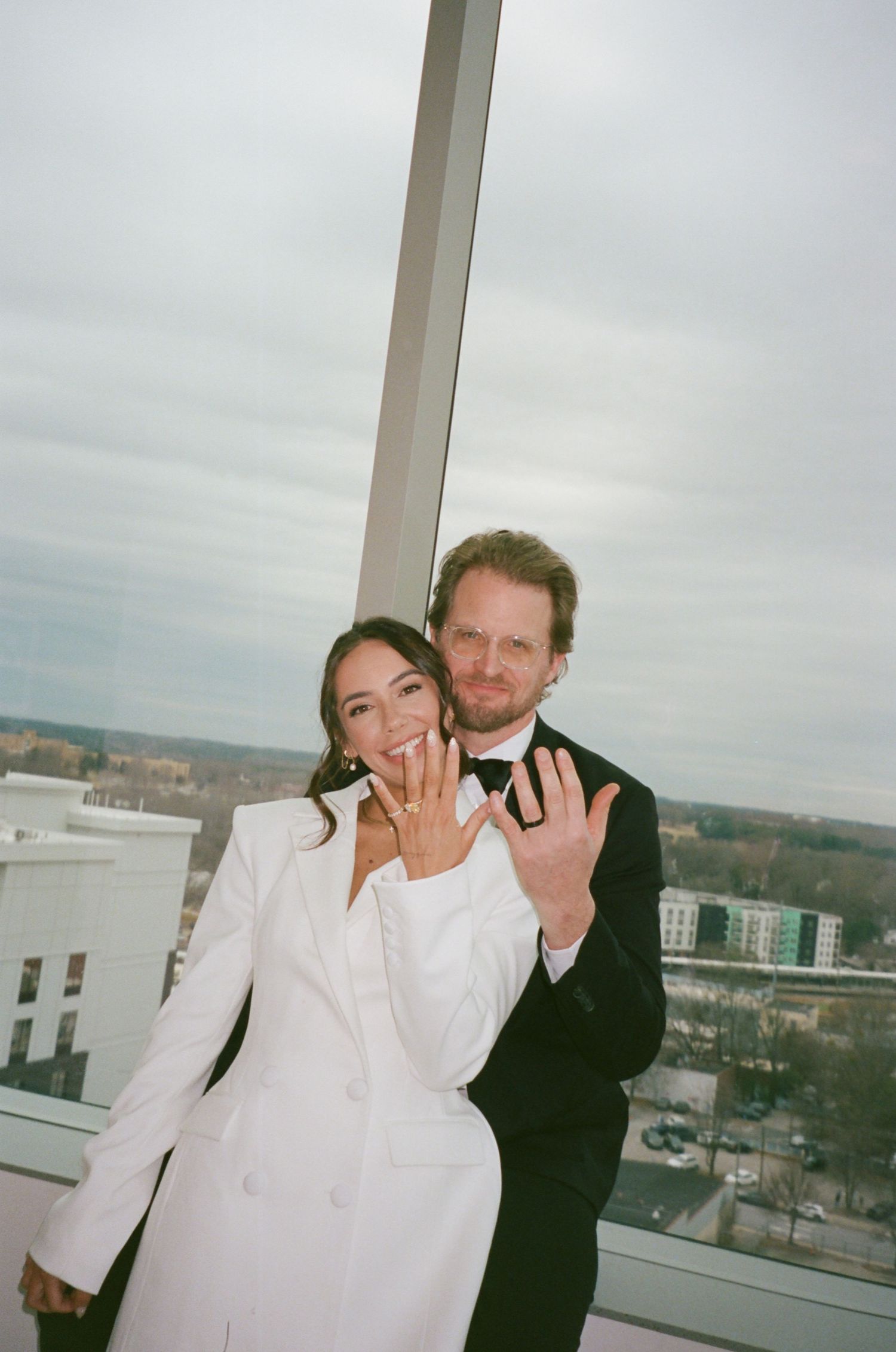 Happy couple in wedding attire showing off rings on a rooftop with city views.
