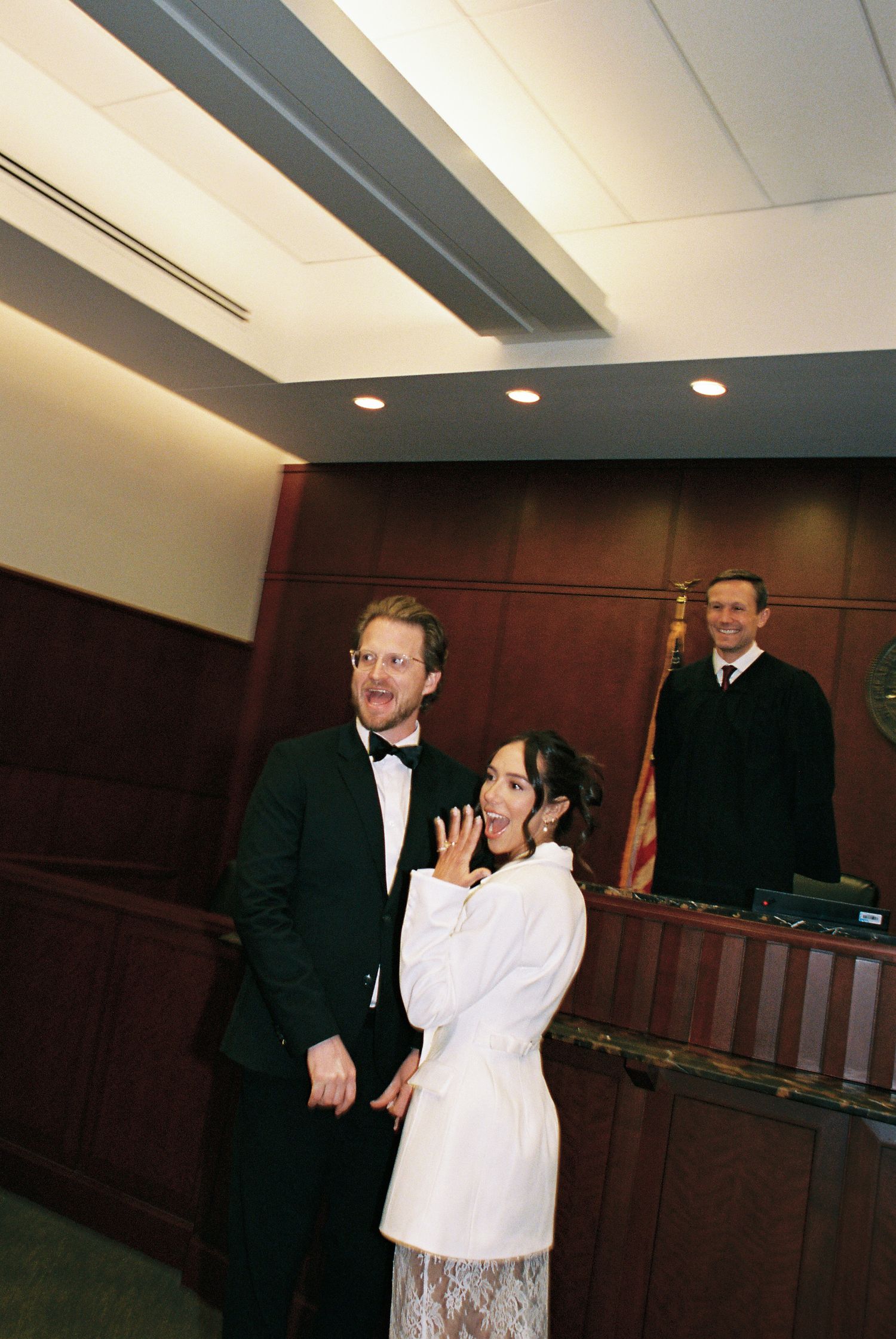 Couple poses in courthouse after civil wedding ceremony, bride shows ring while judge stands at bench behind them.