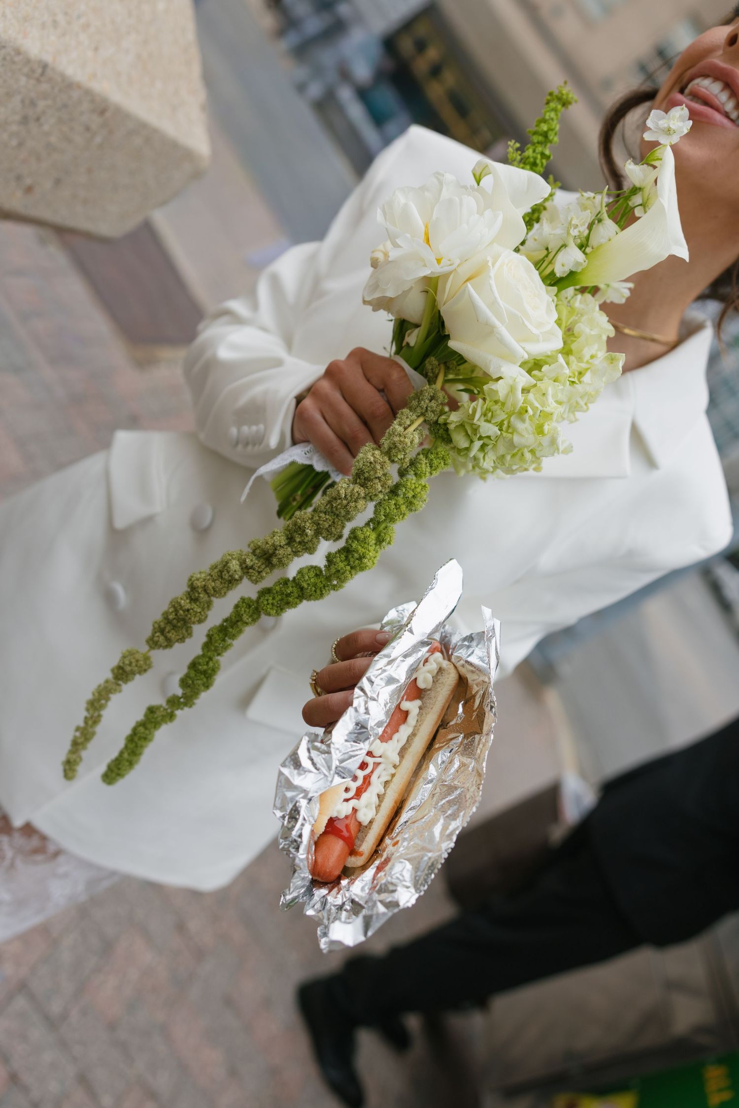 Bride in white wedding dress holding bouquet of white roses and green flowers while eating a hot dog wrapped in foil.