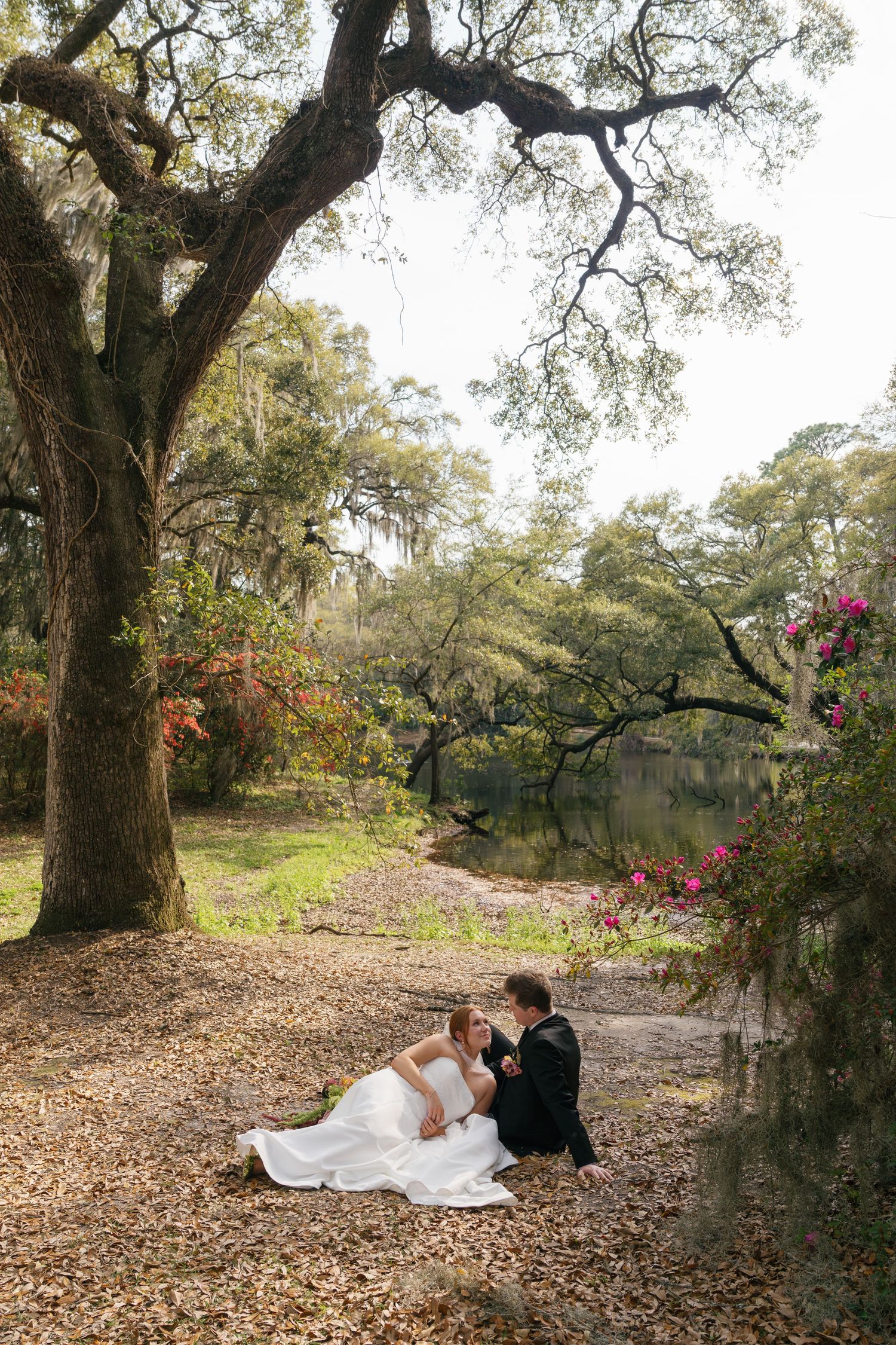 Bride and groom sitting together on fallen leaves beside a tranquil lake under a large oak tree with Spanish moss.