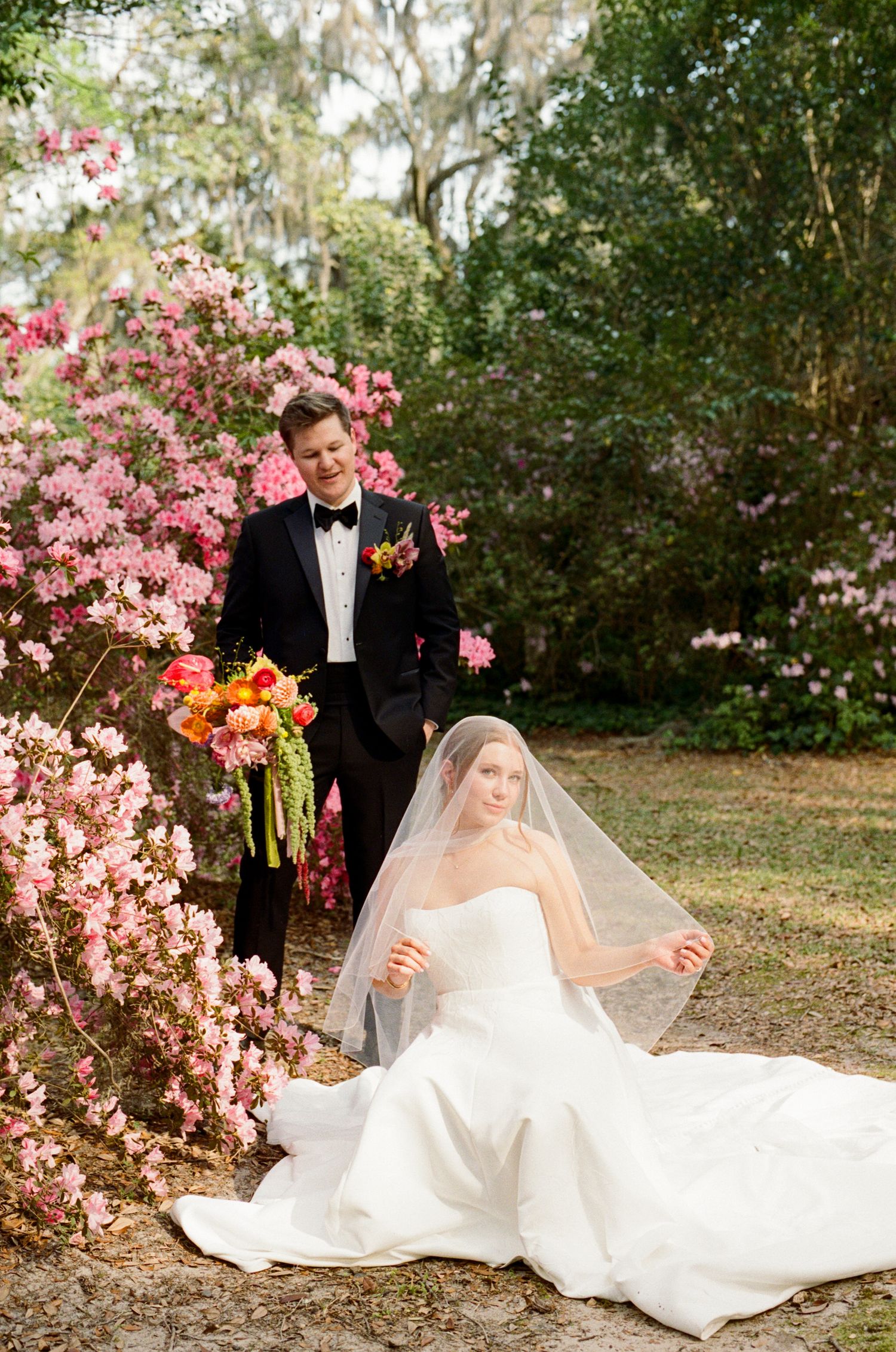 Bride in white gown with veil sits outdoors while groom in black tuxedo holds colorful bouquet near pink azalea bushes.