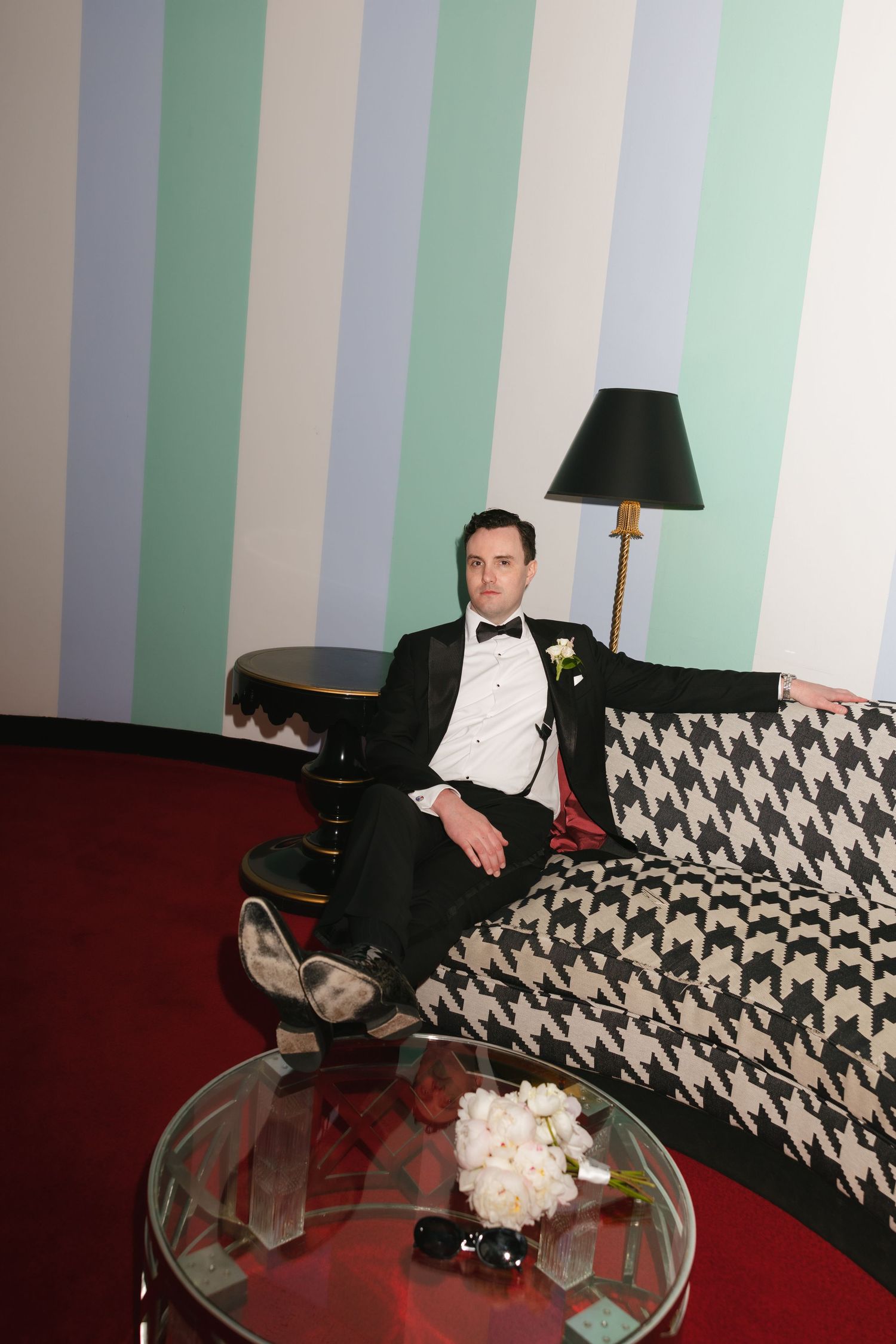 A man in a tuxedo with boutonniere sits on a houndstooth sofa beside a lamp and striped wall.