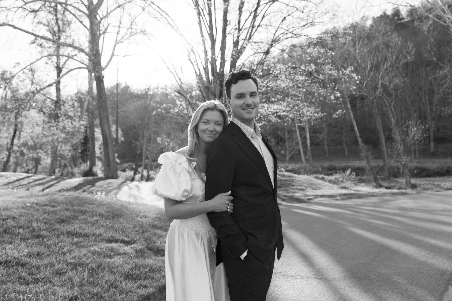 Black and white photo of a smiling couple posing outdoors, woman in white dress and man in dark suit, trees in background.
