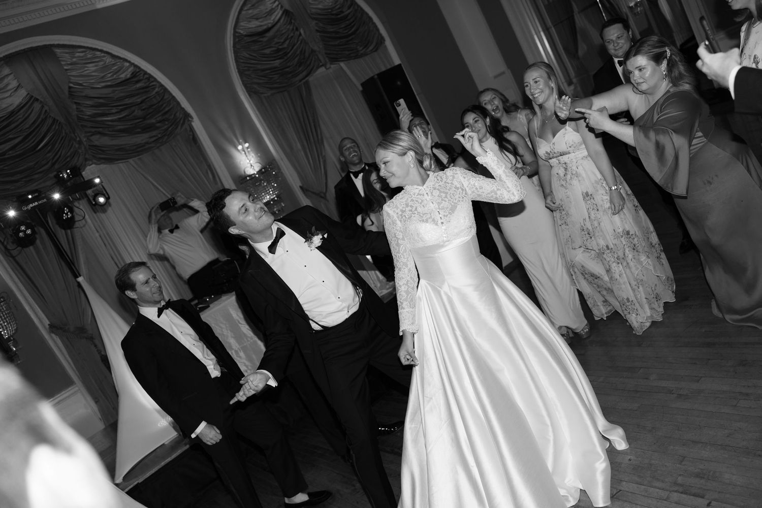 Black and white photo of bride and groom dancing at elegant wedding reception with guests in formal attire.