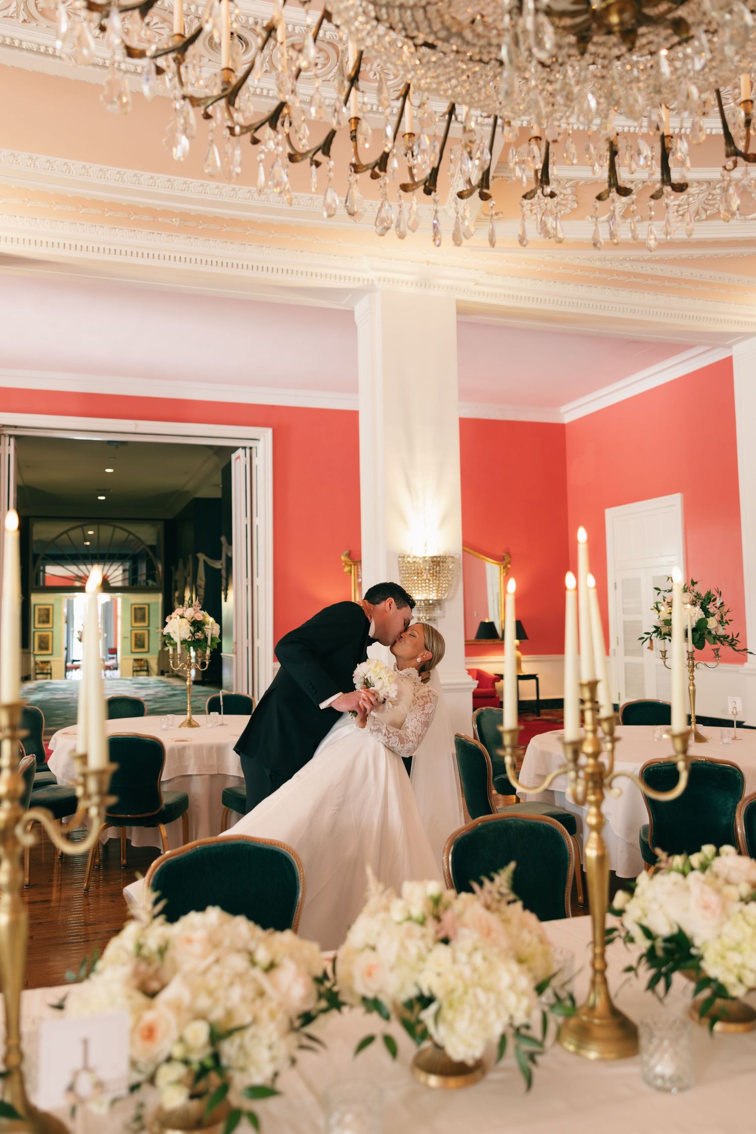 Bride and groom share a romantic kiss in an elegant ballroom with coral walls, crystal chandeliers, and floral centerpieces.