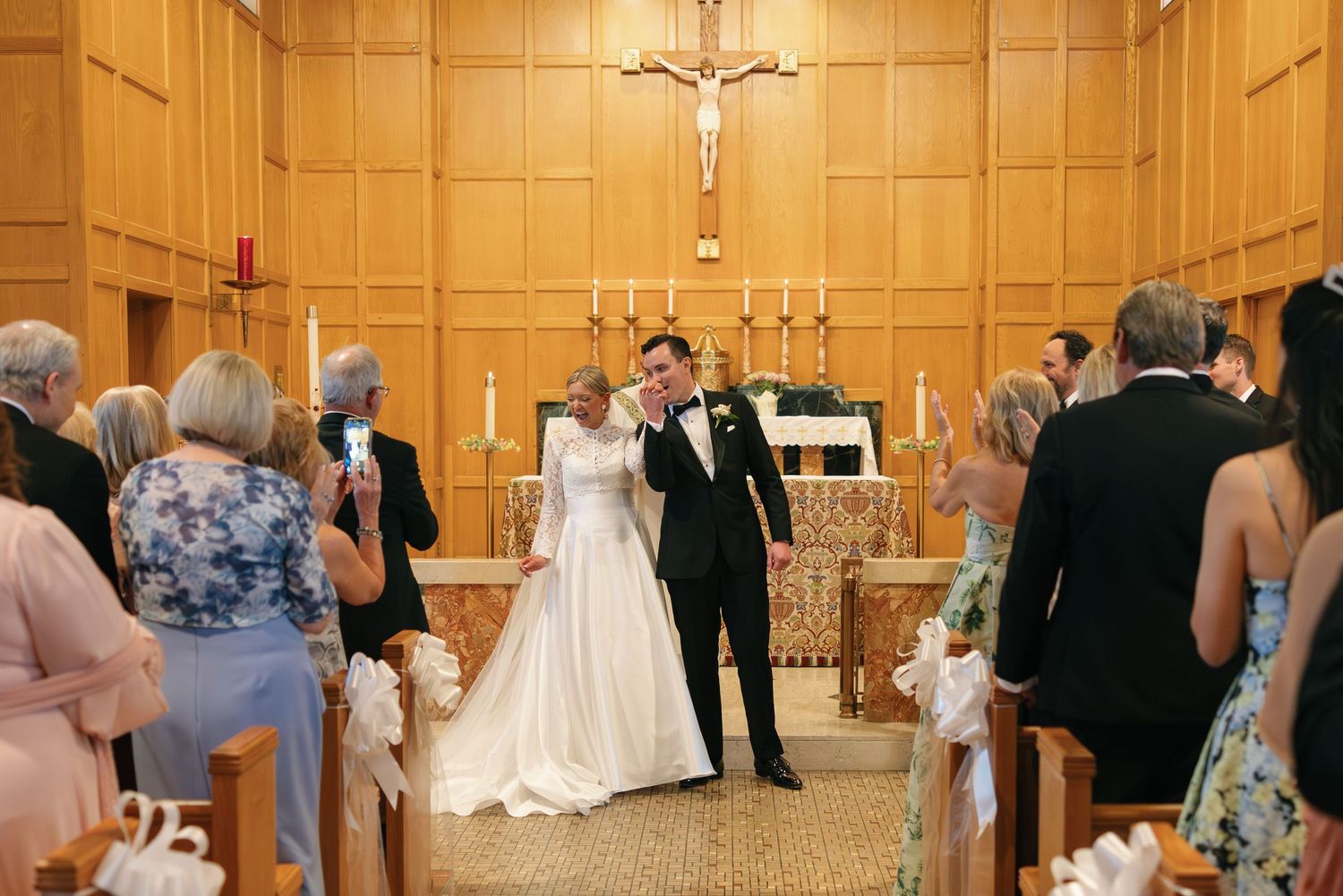 Bride and groom walk down the aisle of a wooden chapel after their Catholic wedding ceremony.