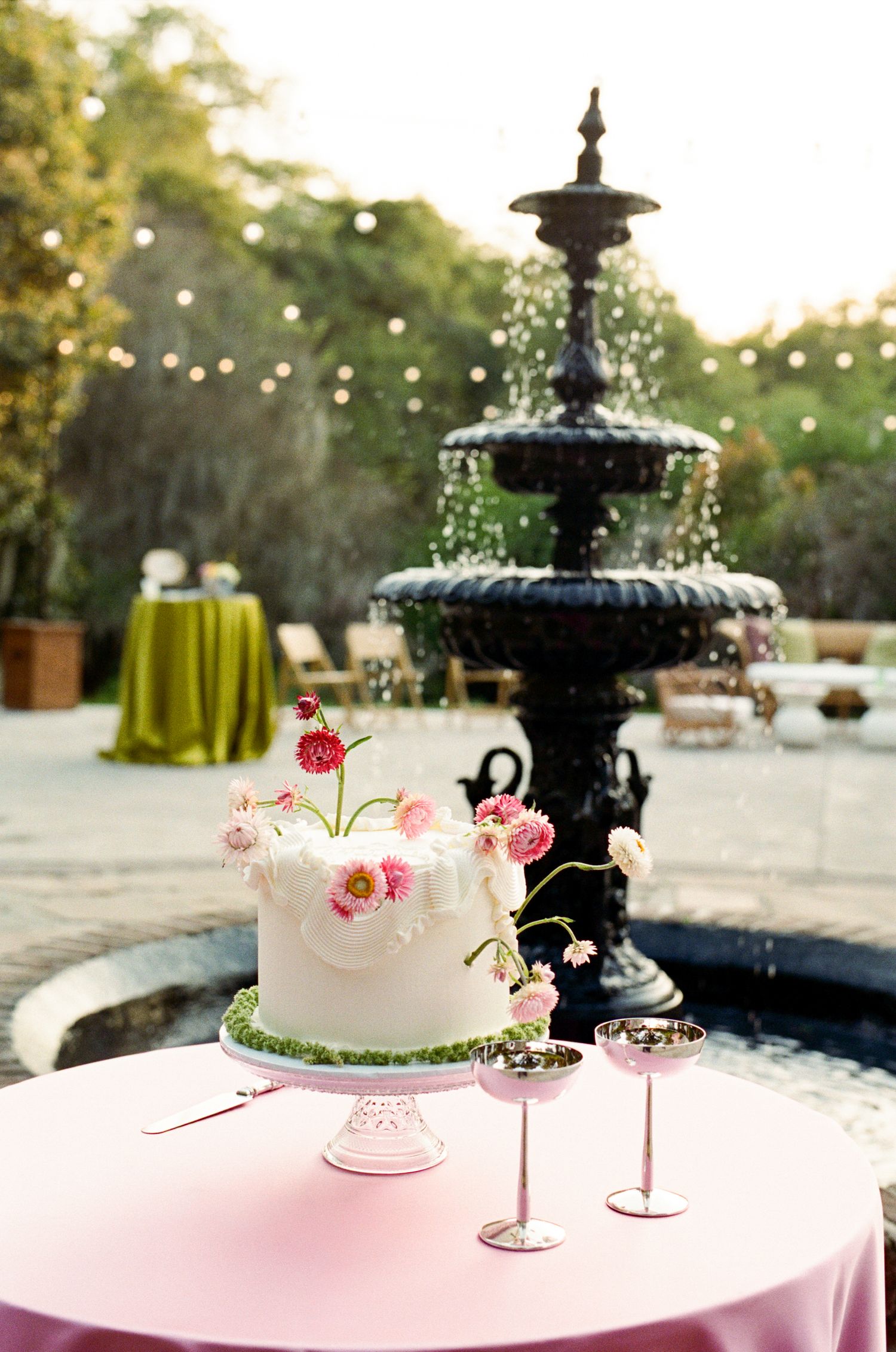 Elegant white wedding cake with pink flowers on a pink table beside a black fountain at an outdoor garden reception.