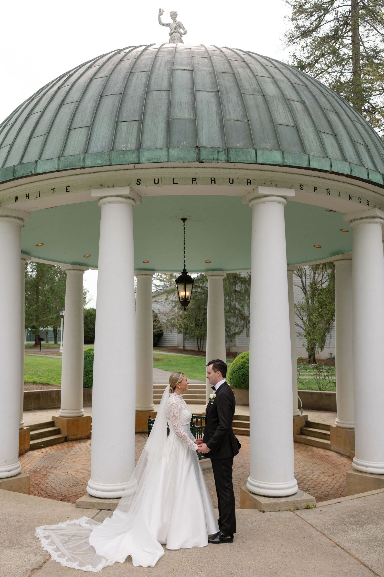 Bride and groom hold hands under the iconic White Sulphur Springs gazebo with white columns and green copper dome.