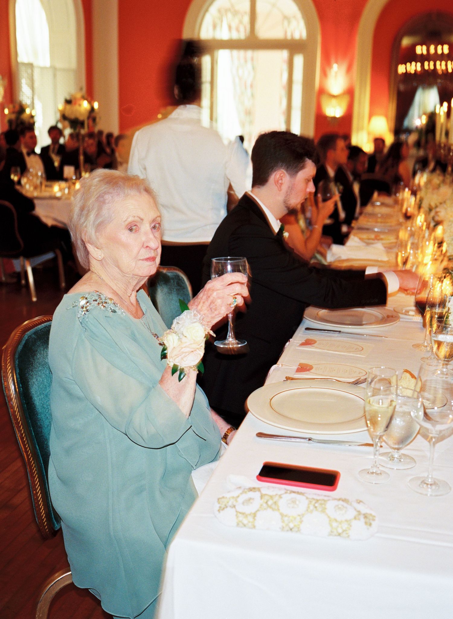 Elderly woman in teal dress holding wine glass at formal wedding reception dinner table.