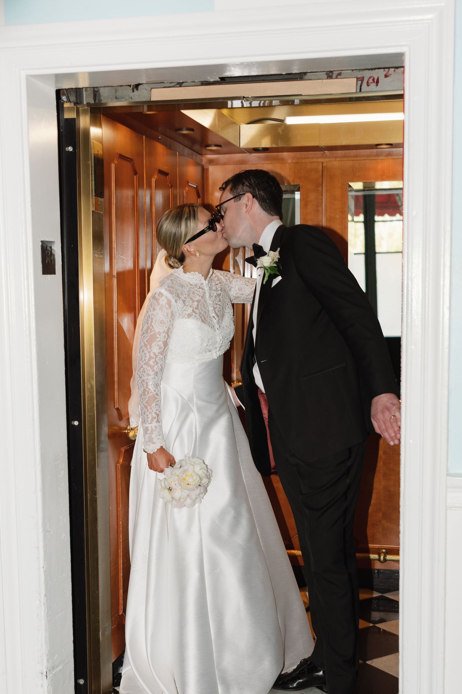 Bride and groom share a romantic kiss in an elegant elevator, bride holding white bouquet in lace wedding gown.