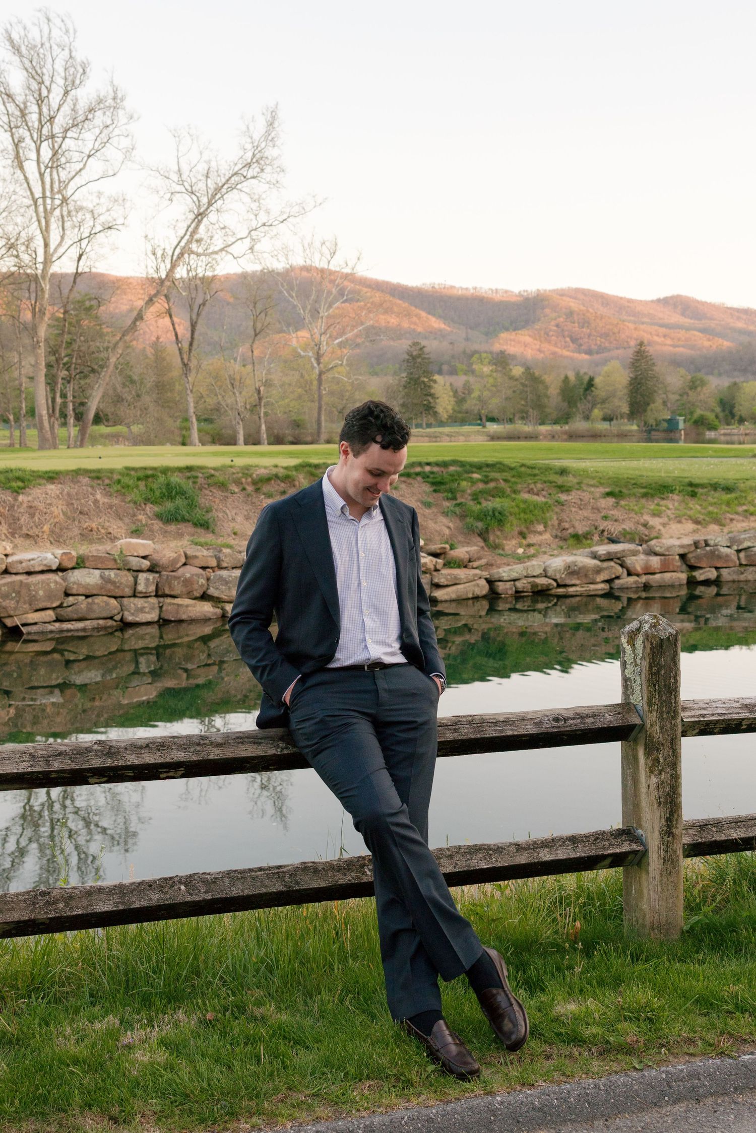 Man in dark suit leaning on wooden fence with mountain landscape and pond in background at sunset.