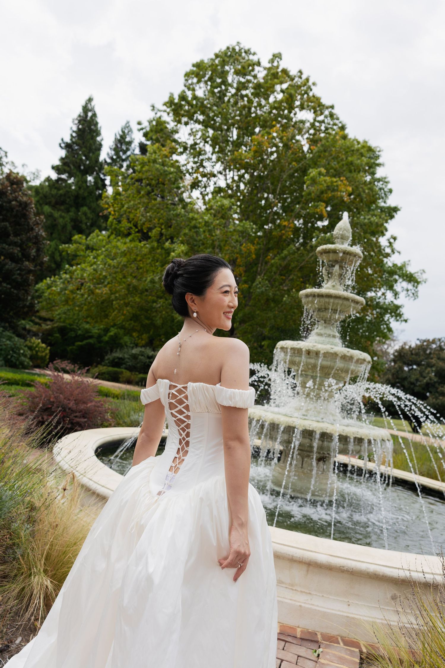 A bride in a white ballgown poses by an ornate tiered fountain in a garden setting during a wedding photoshoot.