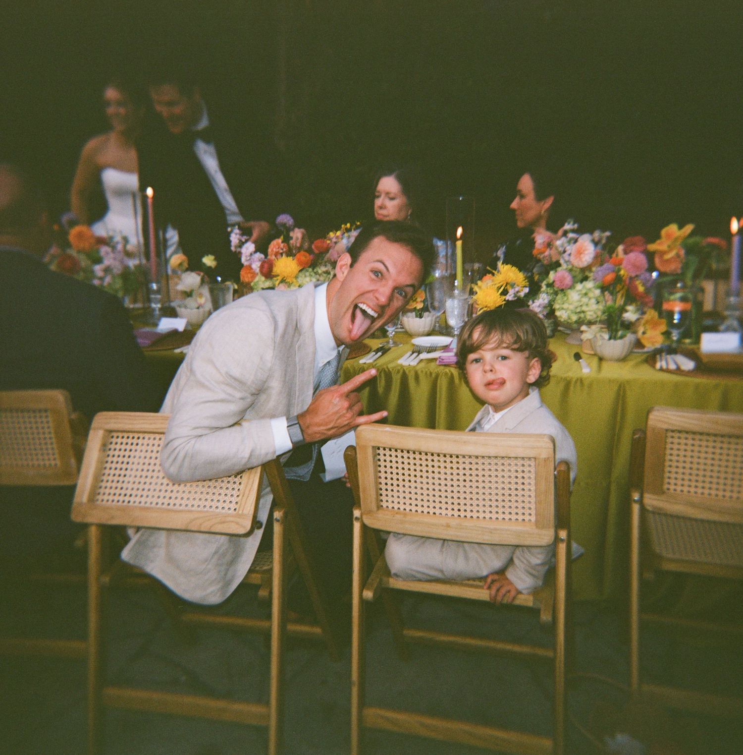 A man and young boy pose playfully at an elegant outdoor wedding reception with colorful floral centerpieces.