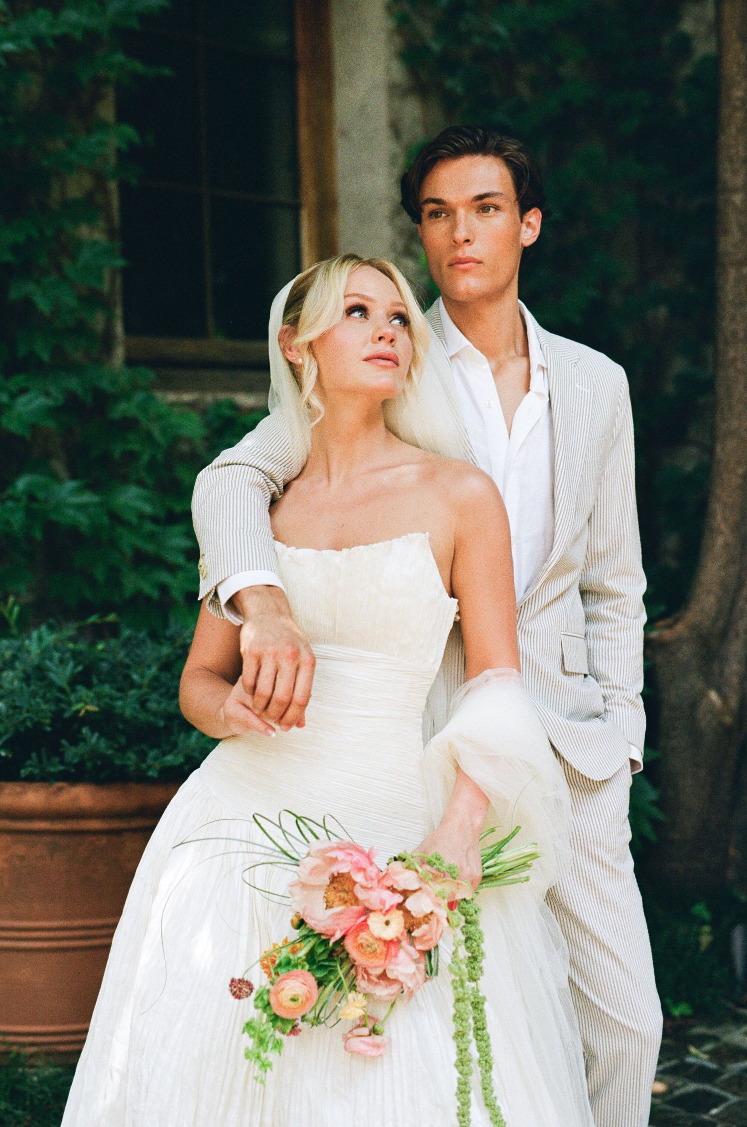 Elegant bride in strapless white gown holding pink bouquet with groom in tan suit posing outdoors near lush greenery.