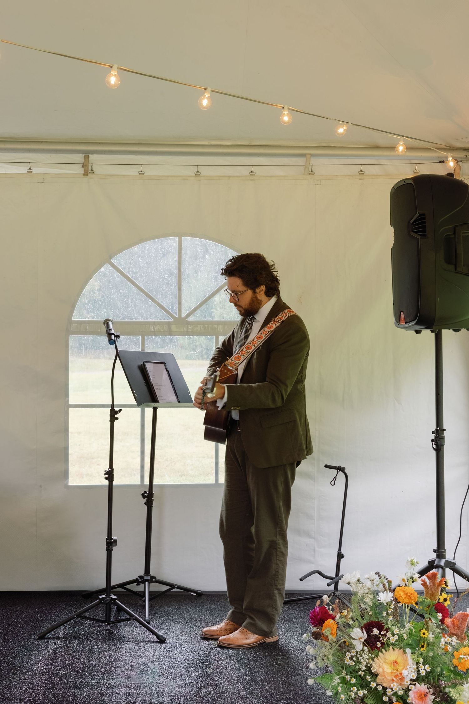 A person stands in a white tent with string lights overhead and colorful flowers arranged at their feet.