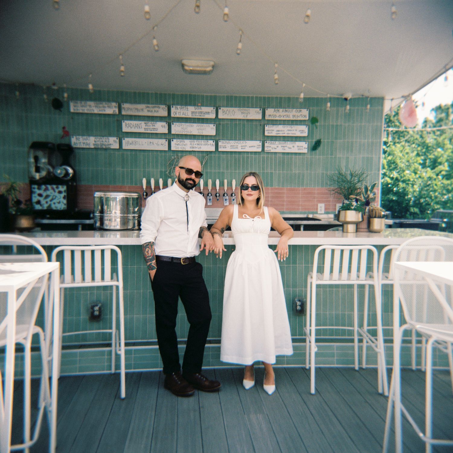 A couple poses at a retro mint green outdoor bar with white barstools and menu boards on the wall.