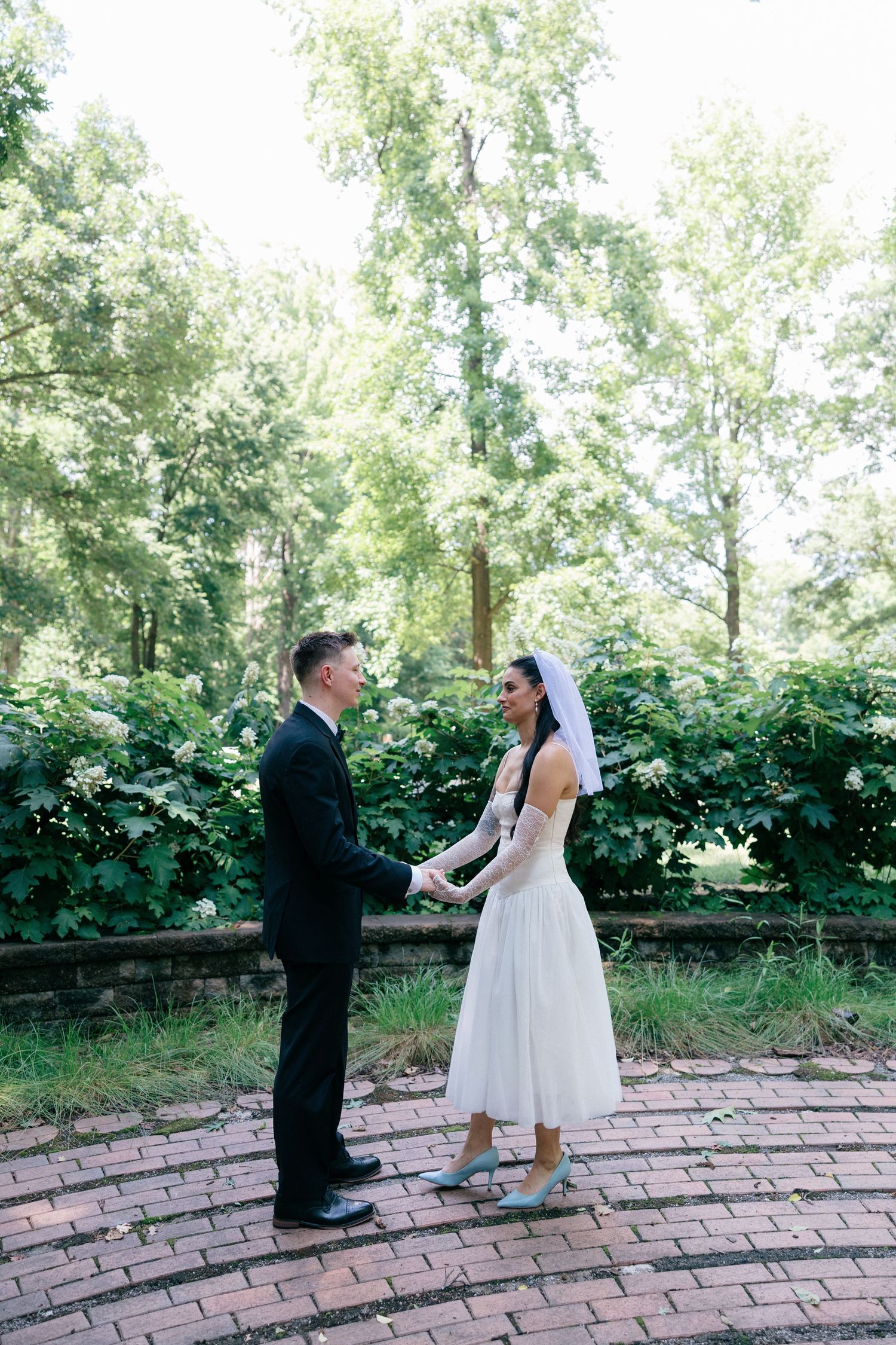 Bride in a short white dress and veil holds hands with groom in a black suit on a brick path surrounded by lush green trees.