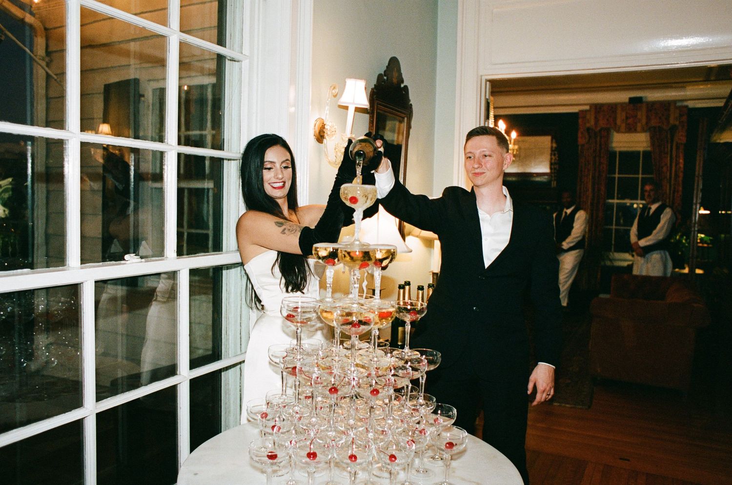 Bride and groom pour champagne together over a tall pyramid of coupe glasses during an elegant wedding reception.