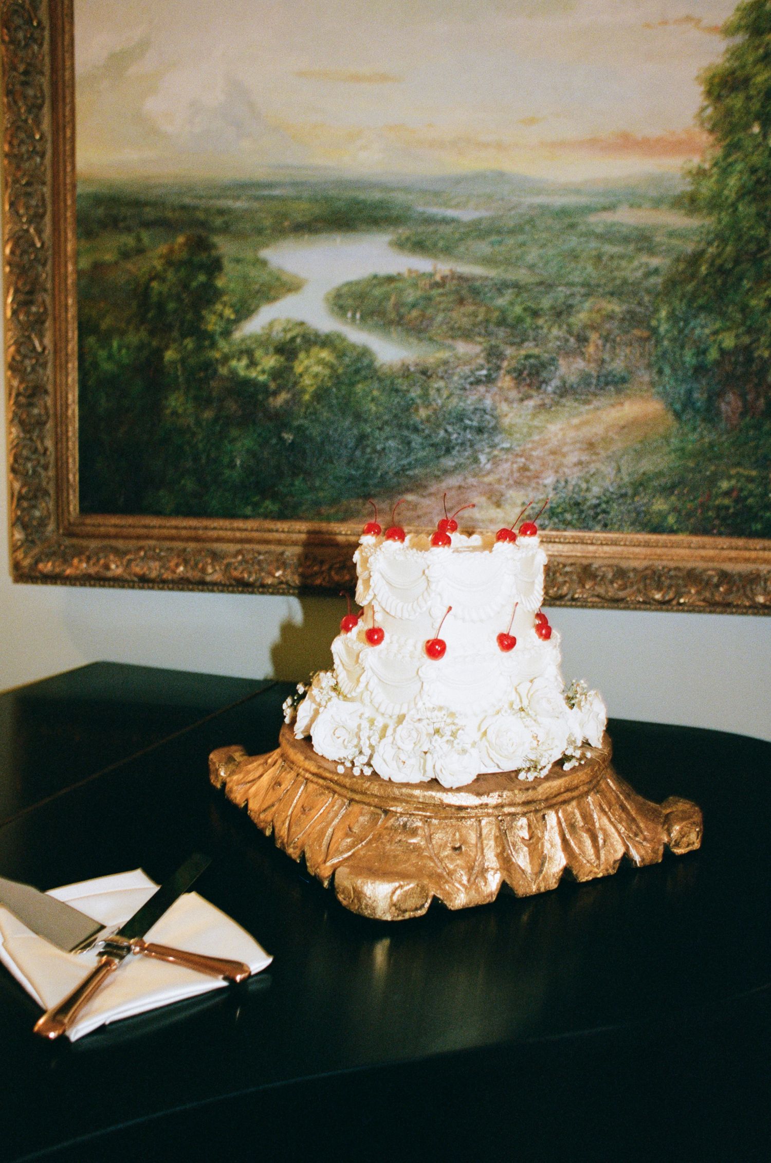 Two-tier white wedding cake with red cherries on an ornate gold stand atop a black piano, beside a cake knife and napkin, below a landscape 