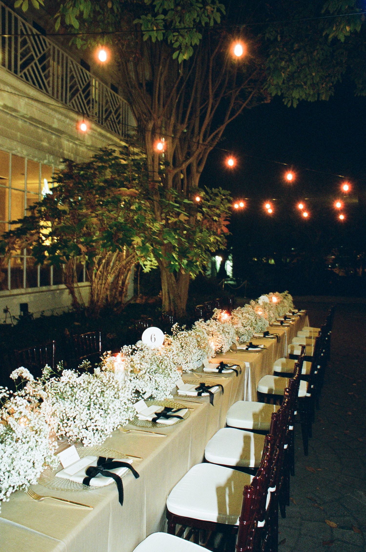 Long outdoor wedding reception table with baby's breath, black ribbon napkins, and string lights glowing at night.
