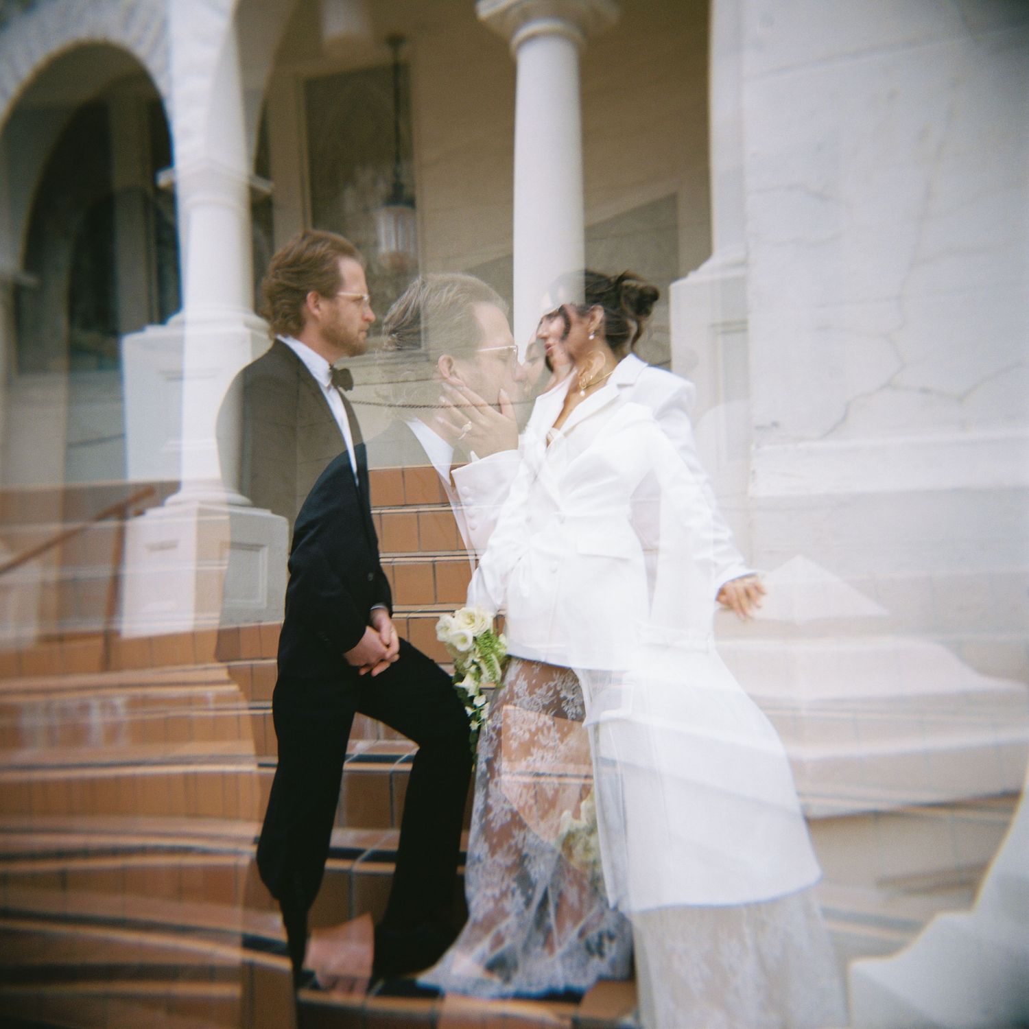 A couple in wedding attire stands on elegant steps with white columns, the bride in a white jacket and groom in a black suit.
