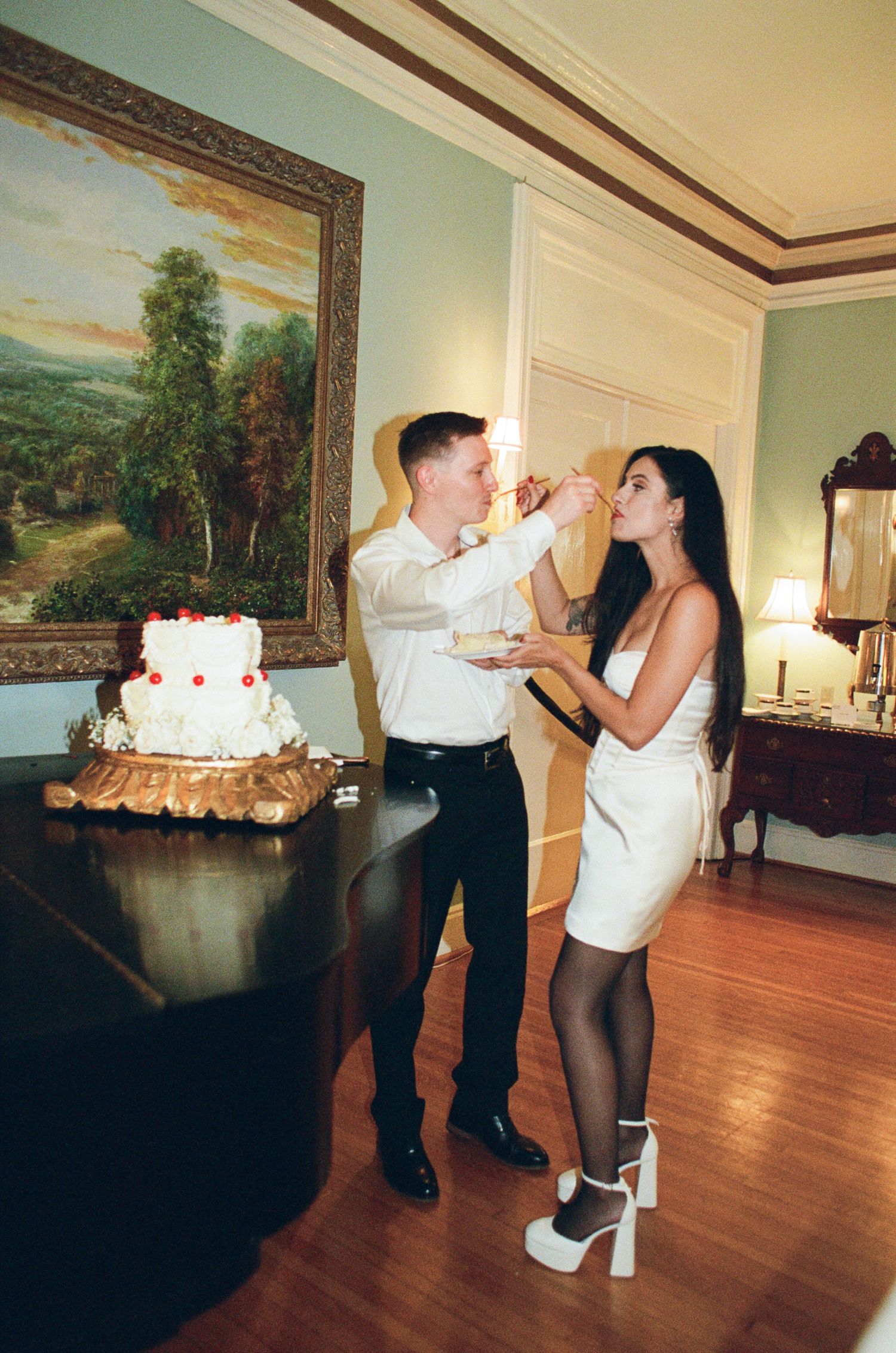 A man in a white shirt and a woman in a white dress feed each other cake beside a white tiered wedding cake on a grand piano.