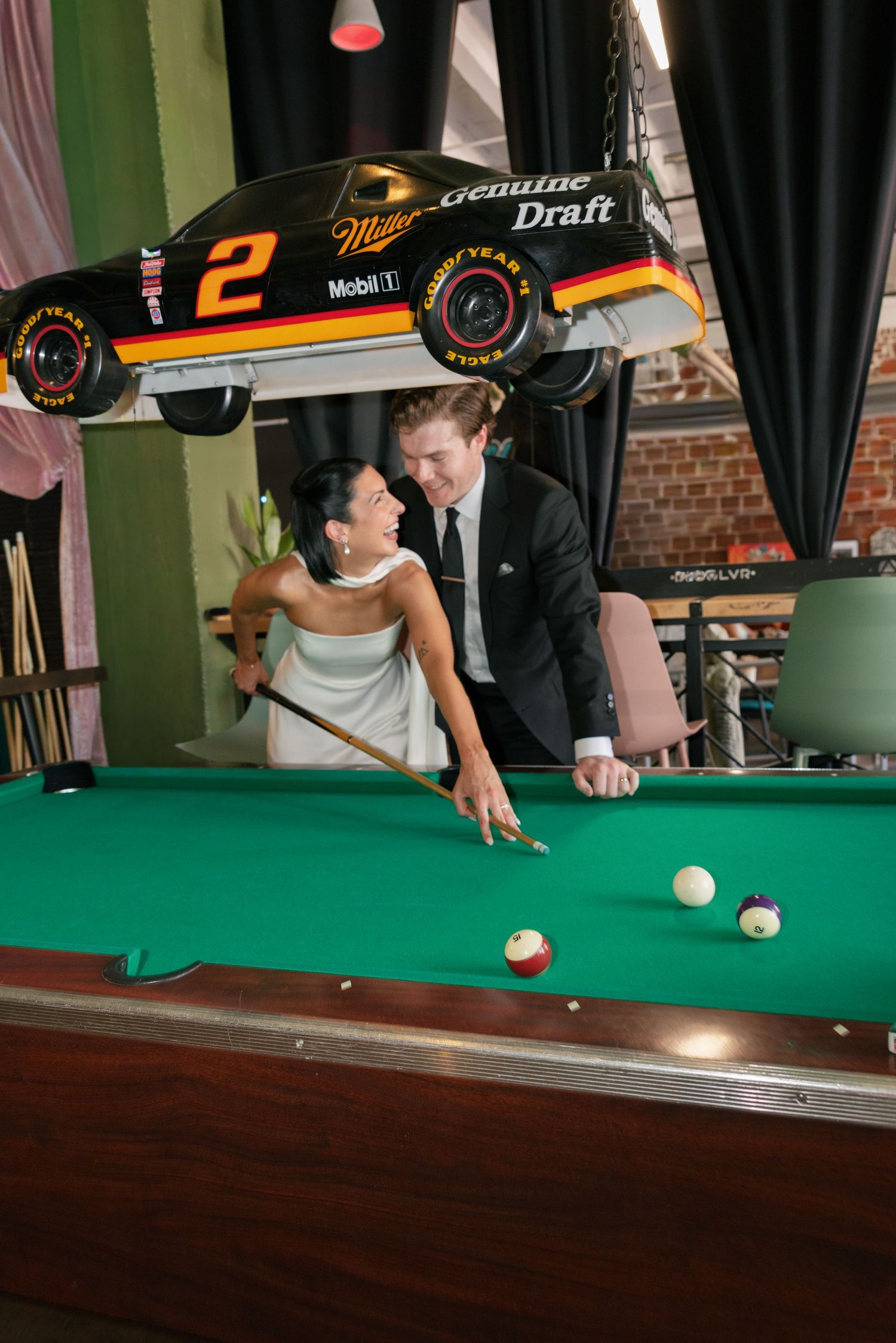 Bride and groom share a laughing moment while playing pool beneath a hanging Miller Genuine Draft race car at a lively venue.