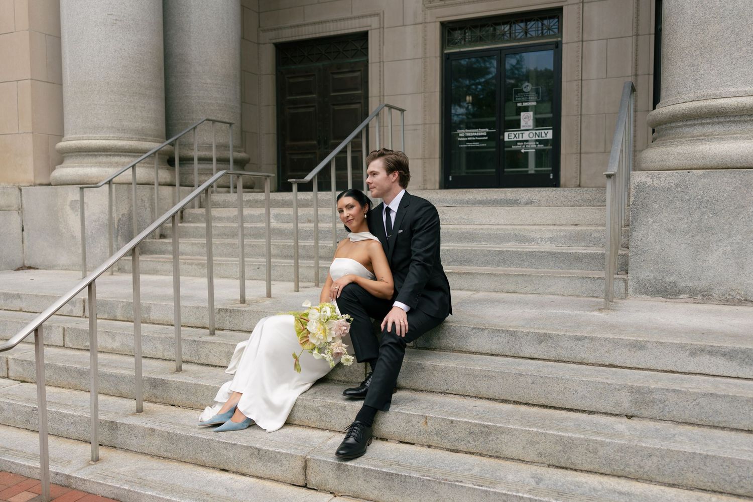 Bride in a white gown holding a bouquet sits beside a groom in a dark suit on the stone steps of a columned building.