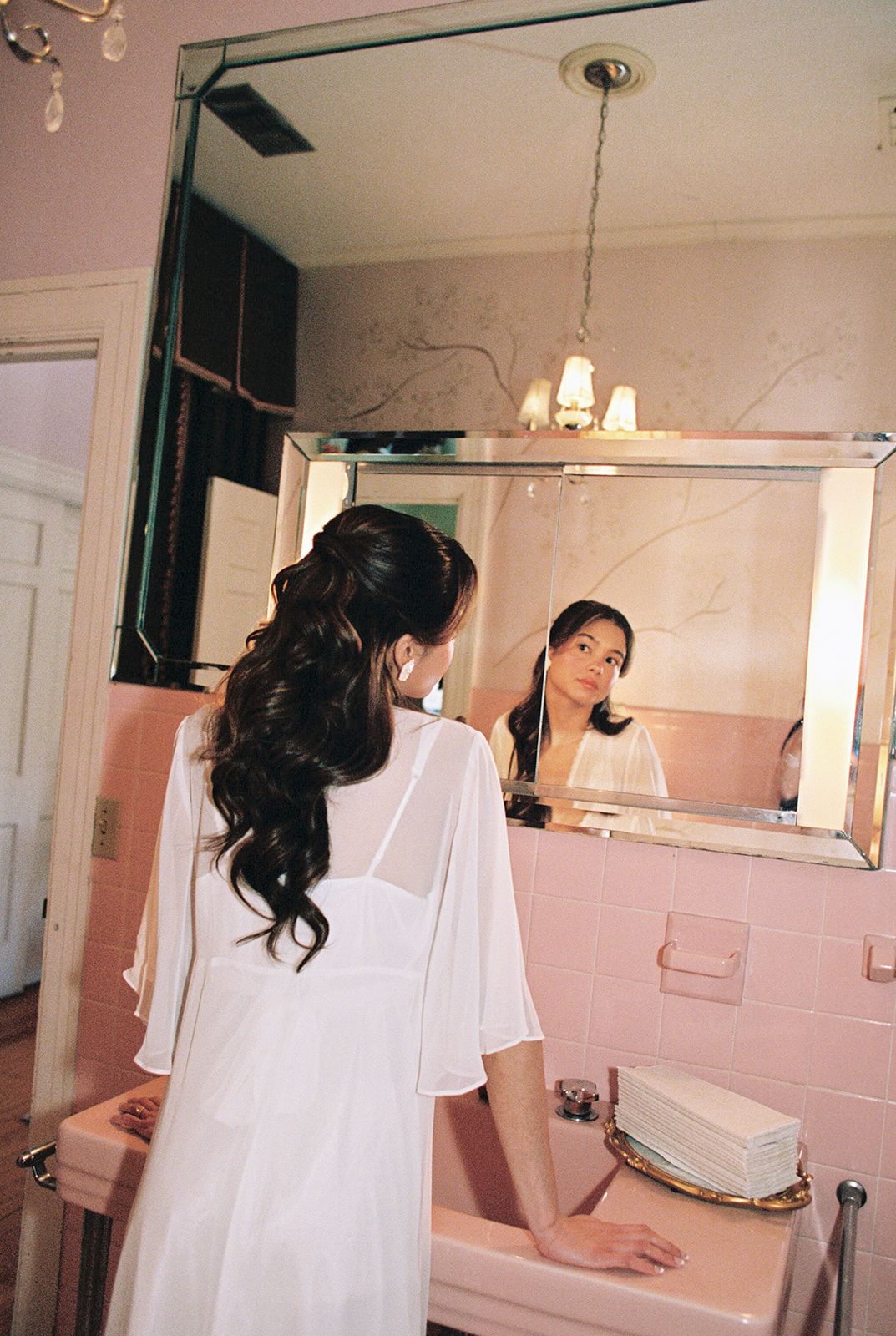 Bride in white robe admiring her elegant half-up hairstyle in a vintage pink bathroom mirror.