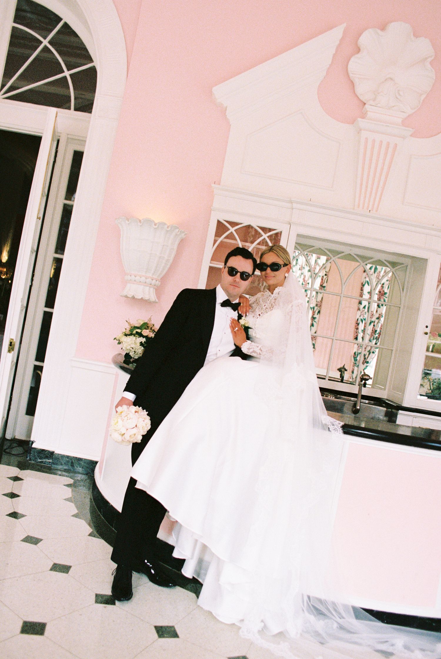 Elegant bride and groom posing together in white wedding dress and black tuxedo inside a pink luxury venue.