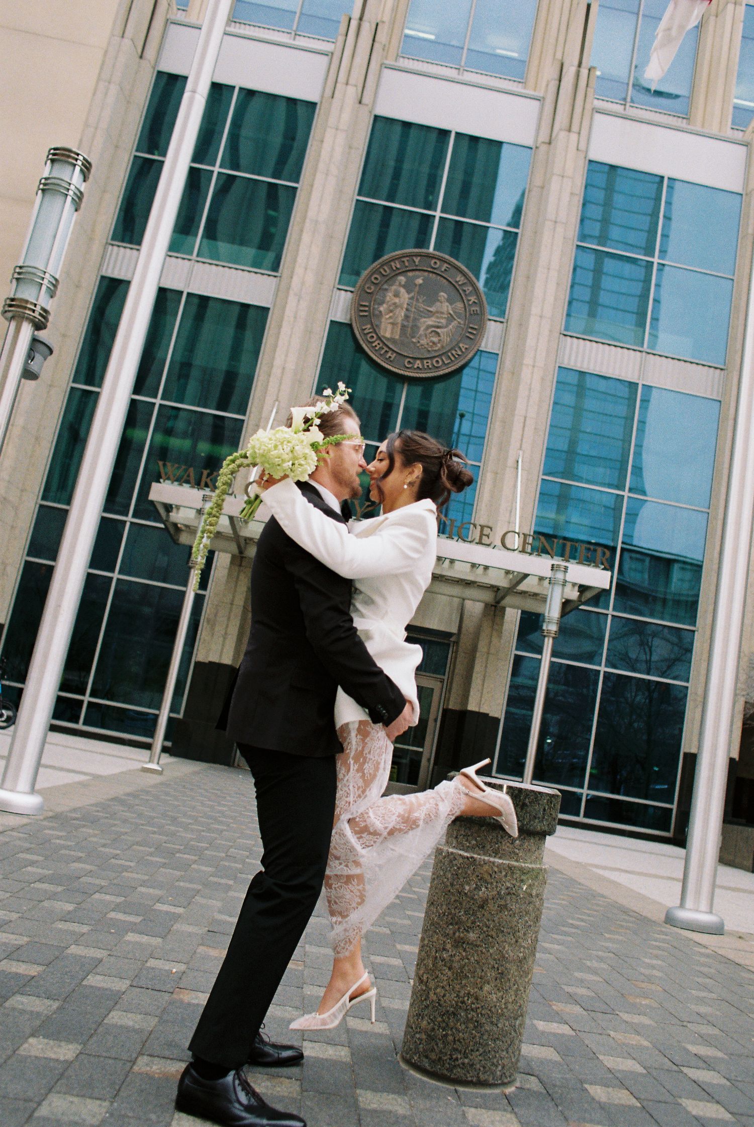Couple sharing a romantic kiss outside a courthouse on their wedding day, bride in lace gown holding bouquet.