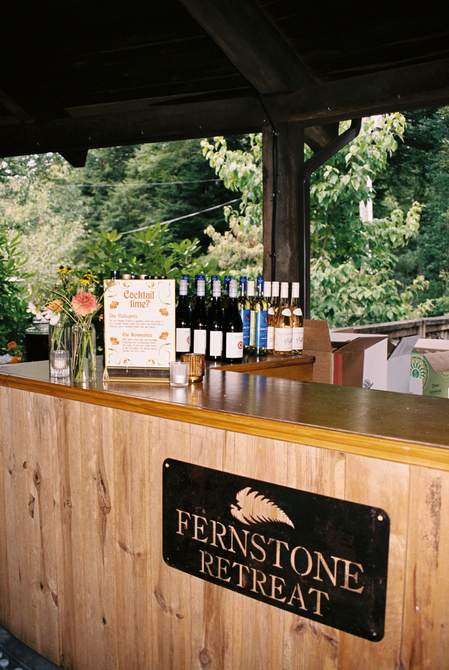 A rustic wooden bar counter with a 'Fernstone Retreat' sign mounted on the side panel displays wine bottles and decor.