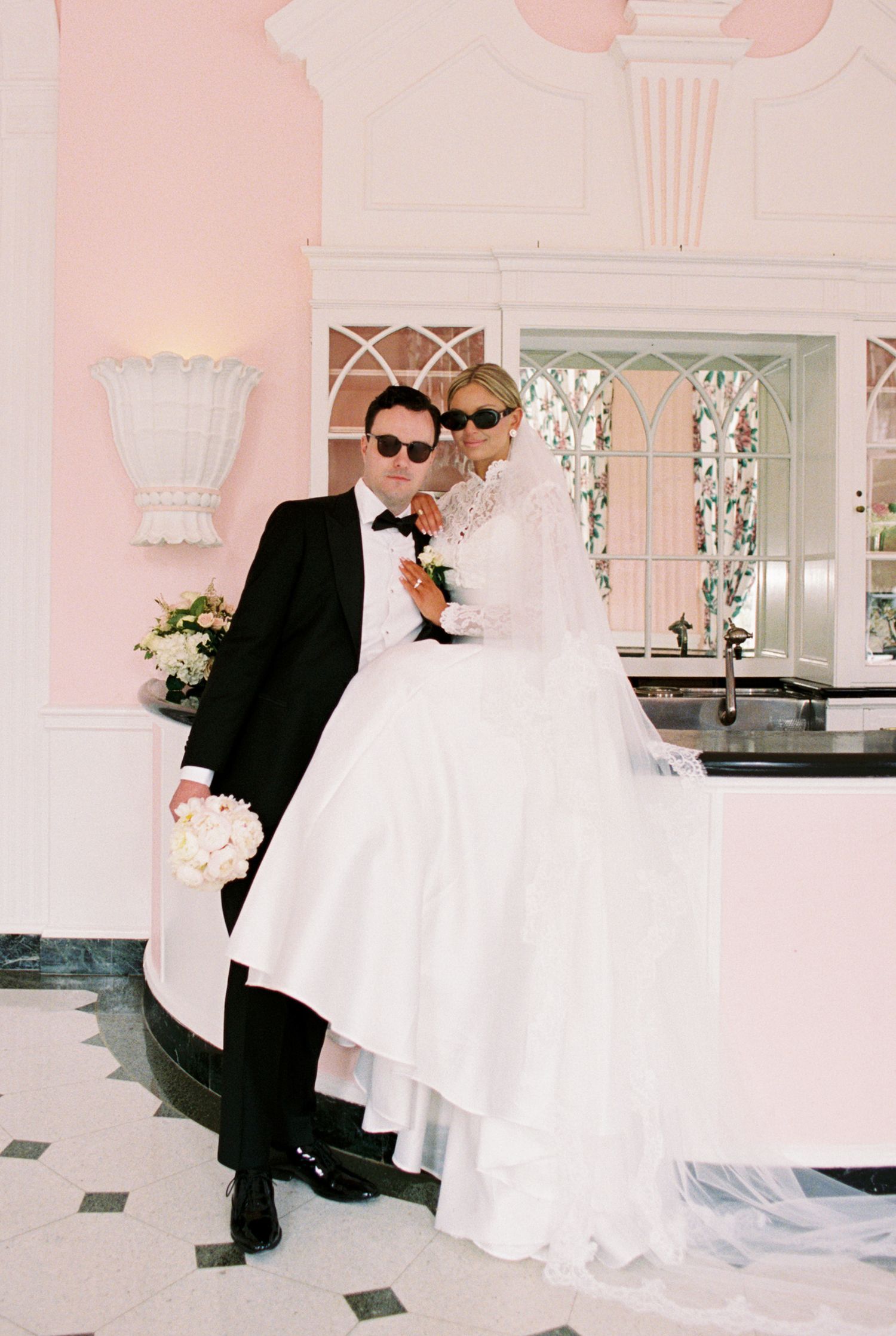 Bride in white gown and groom in black tuxedo pose together wearing sunglasses in an elegant pink venue.