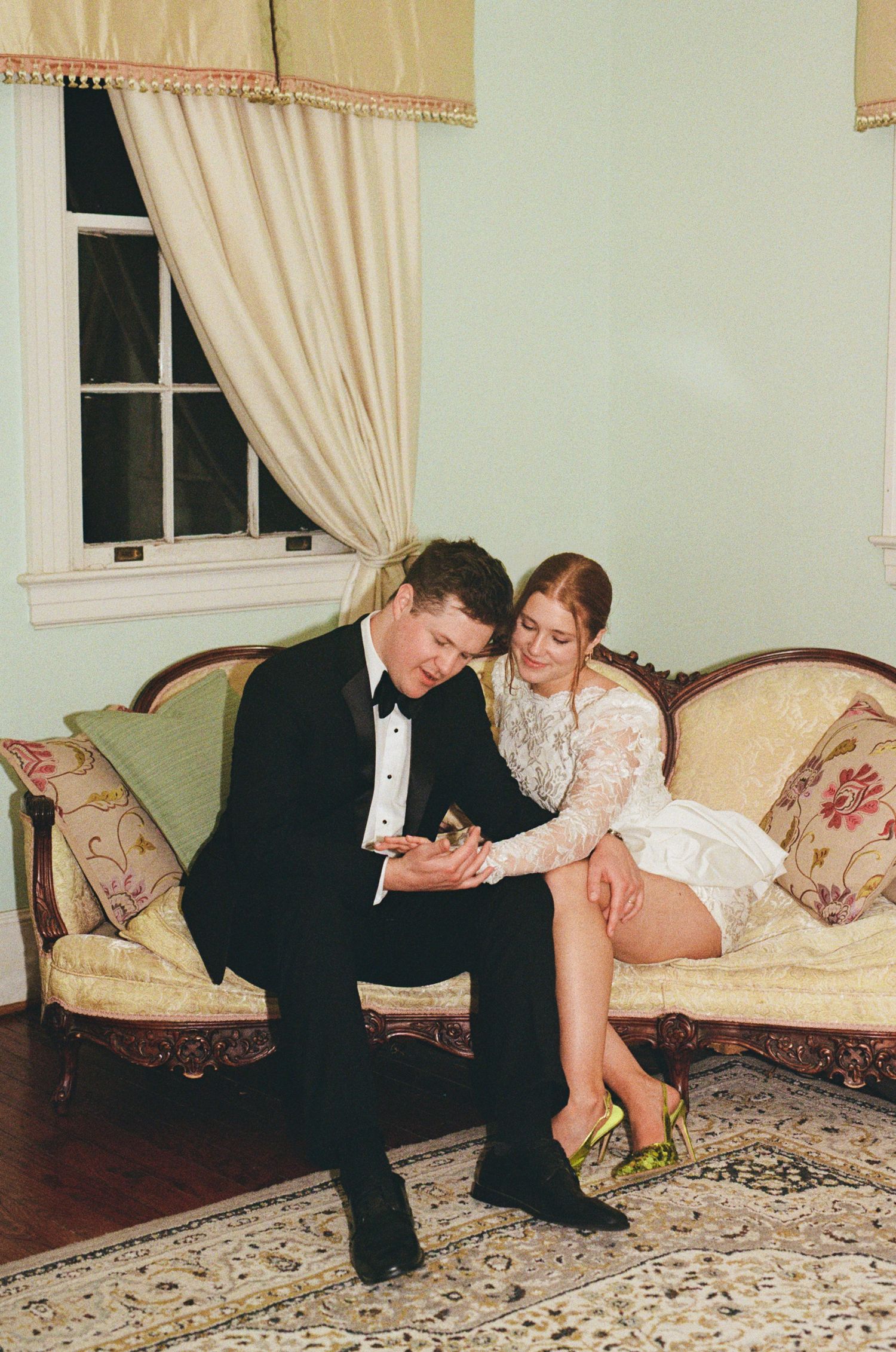 Couple in wedding attire sitting on vintage sofa, groom in tuxedo and bride in white mini dress with gold heels.
