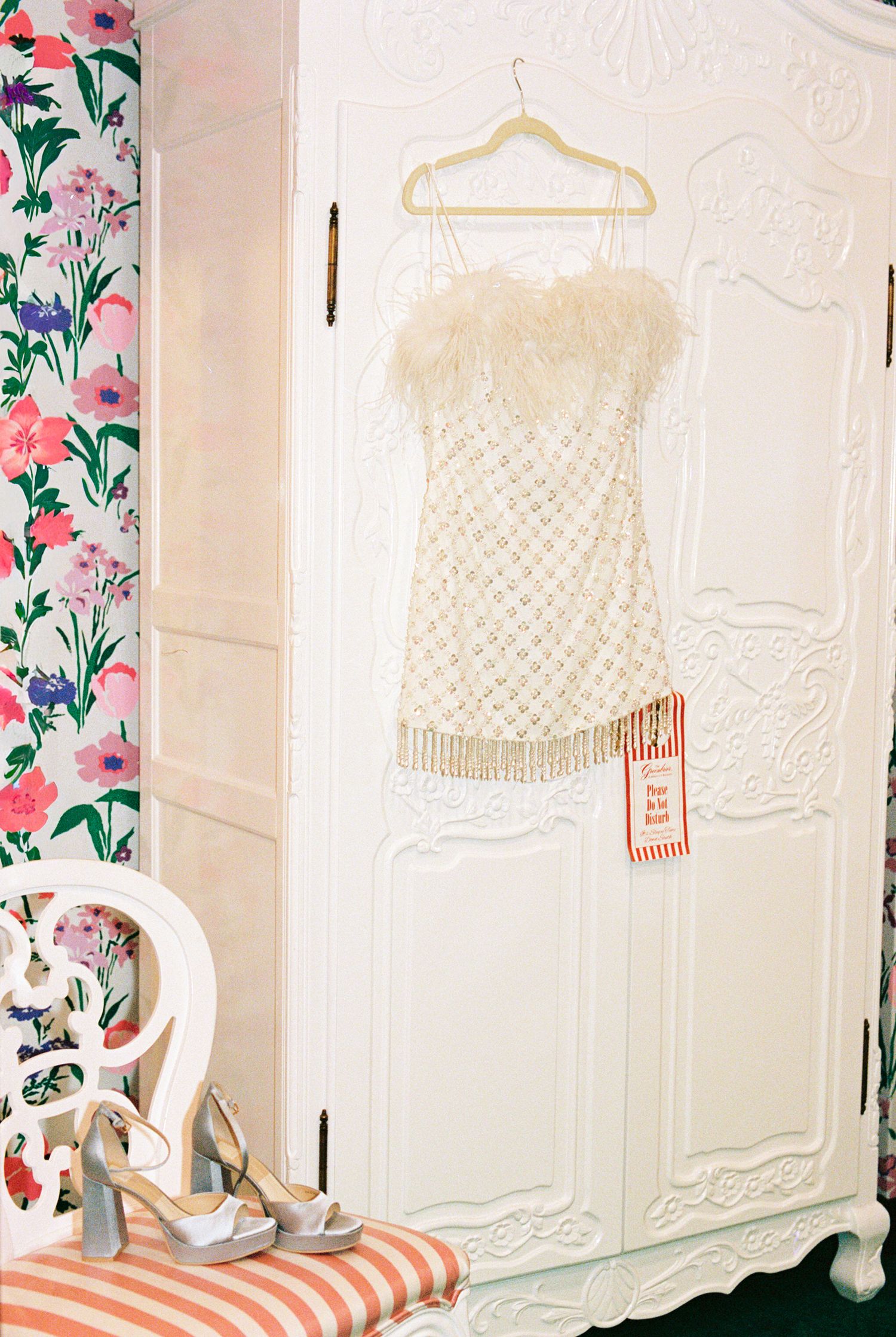 White feather-trimmed mini dress on hanger displayed on ornate white armoire with floral wallpaper backdrop.