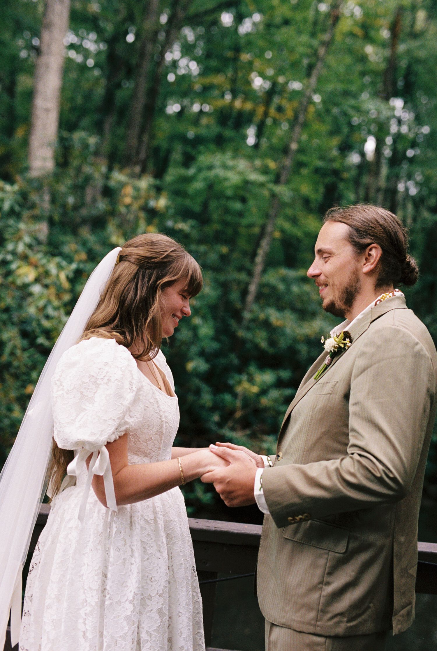 A couple shares a joyful moment during their outdoor forest wedding ceremony, wearing white dress and tan suit.