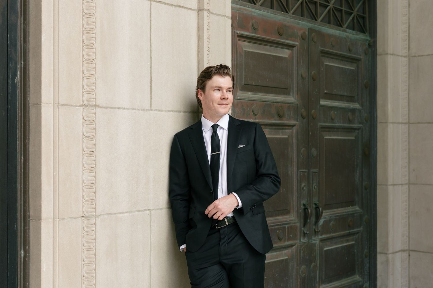 Young man in a black suit and tie leaning casually against a limestone wall beside a large ornate bronze door.