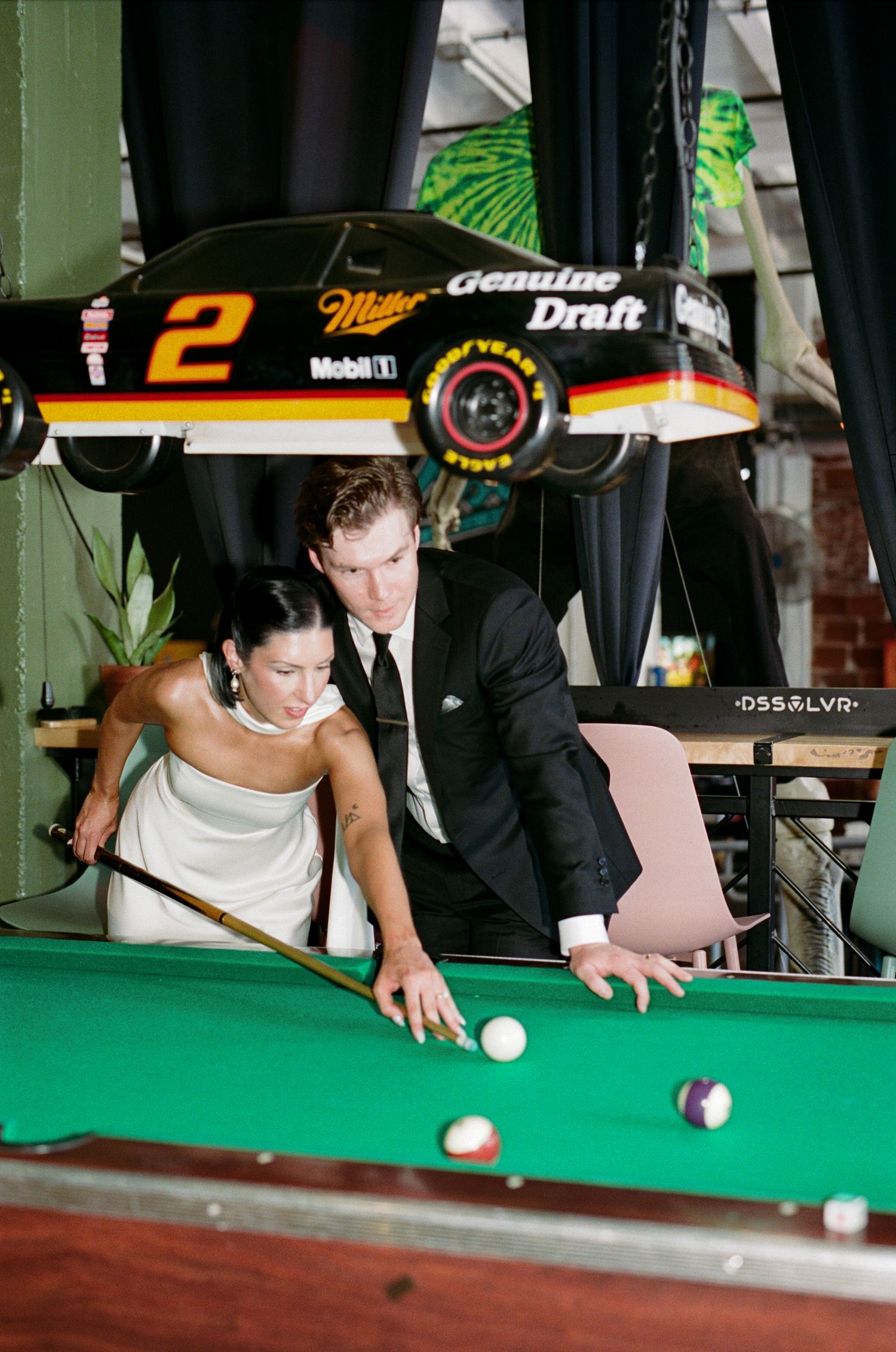 Bride in a white gown and groom in a black suit play pool together beneath a Miller Genuine Draft number 2 race car mounted above the table.
