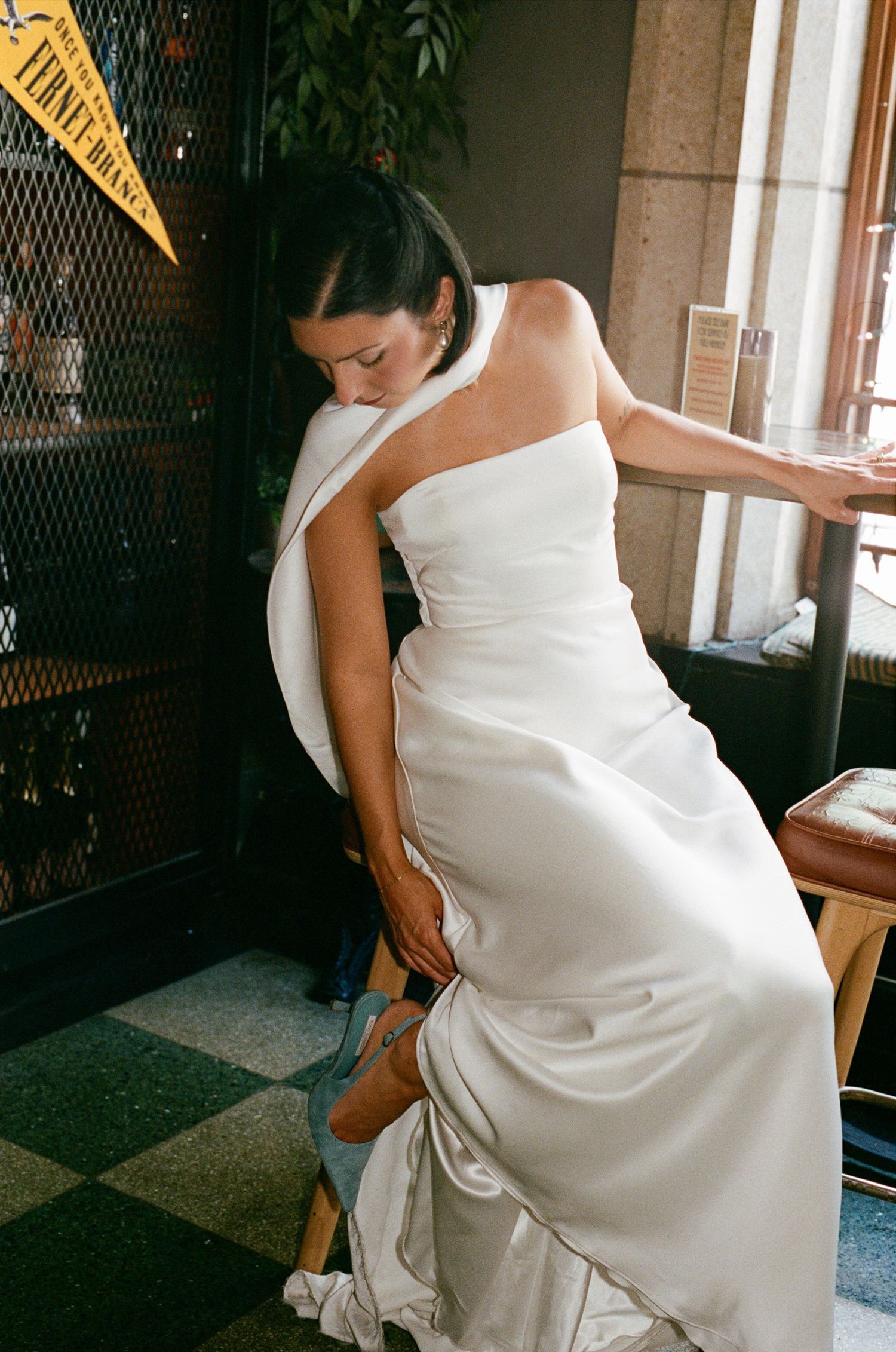 Bride in a white strapless satin wedding gown with a wrap, sitting on a wooden stool and adjusting her shoe in a tiled restaurant.