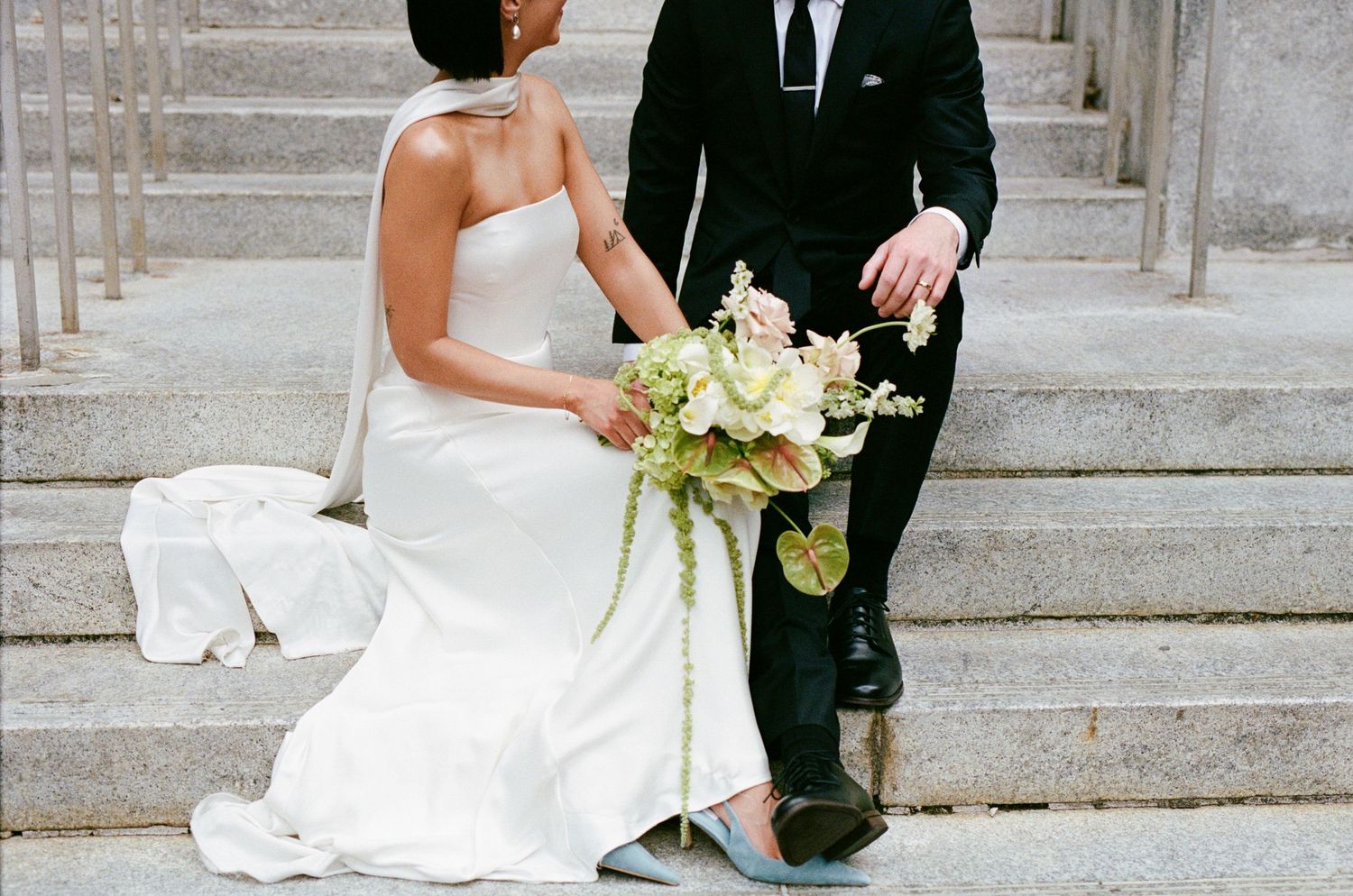 Bride in white gown and groom in black suit sit on stone steps holding a lush green and white wedding bouquet.