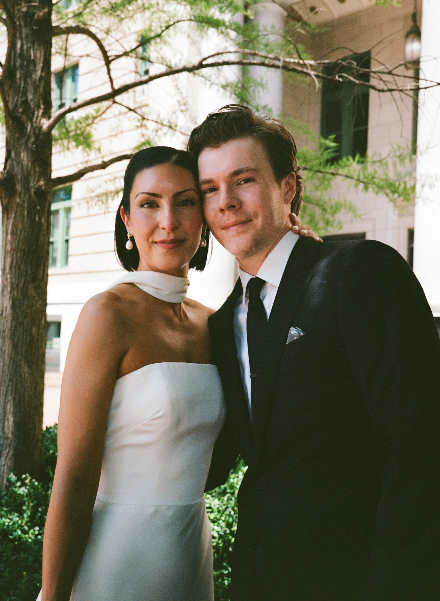 A smiling couple poses outdoors, she in a white strapless dress with a choker collar and he in a black suit and tie.