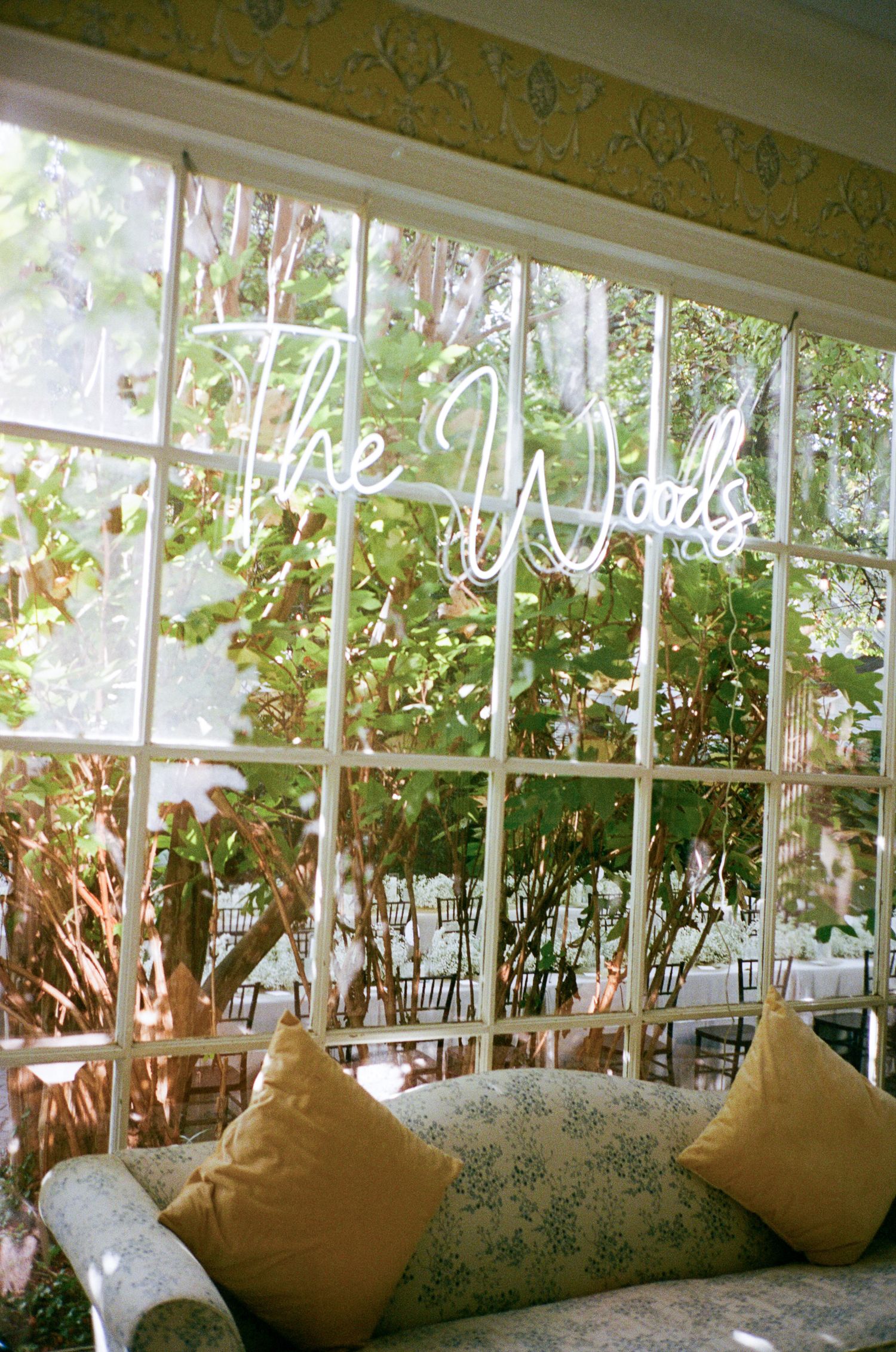 Neon sign reading The Woods in a multi-pane window overlooking a lush garden, with a floral sofa and mustard yellow pillows.