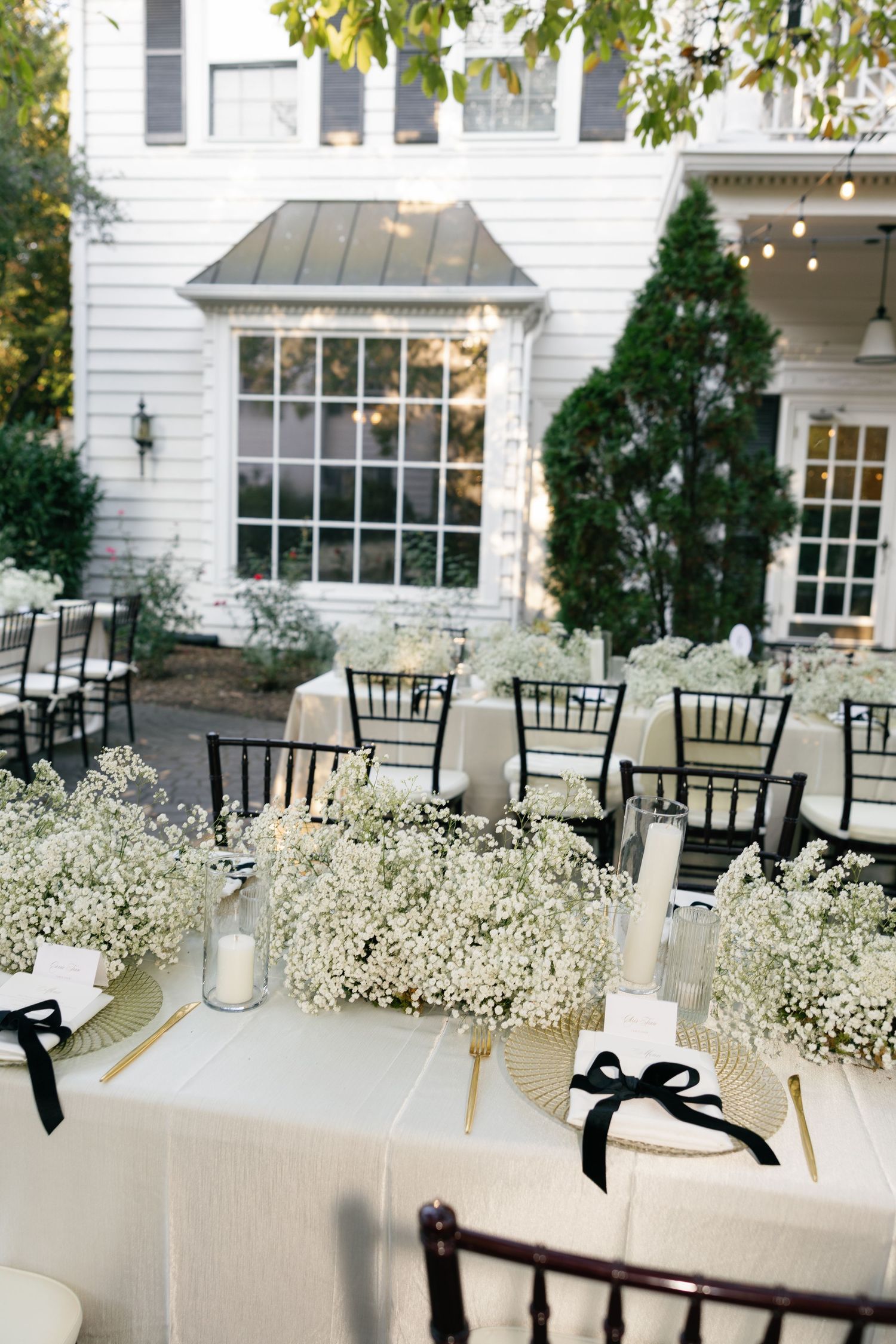 Elegant outdoor wedding reception tables with baby's breath centerpieces, black ribbon napkins, and black chairs beside a white house.