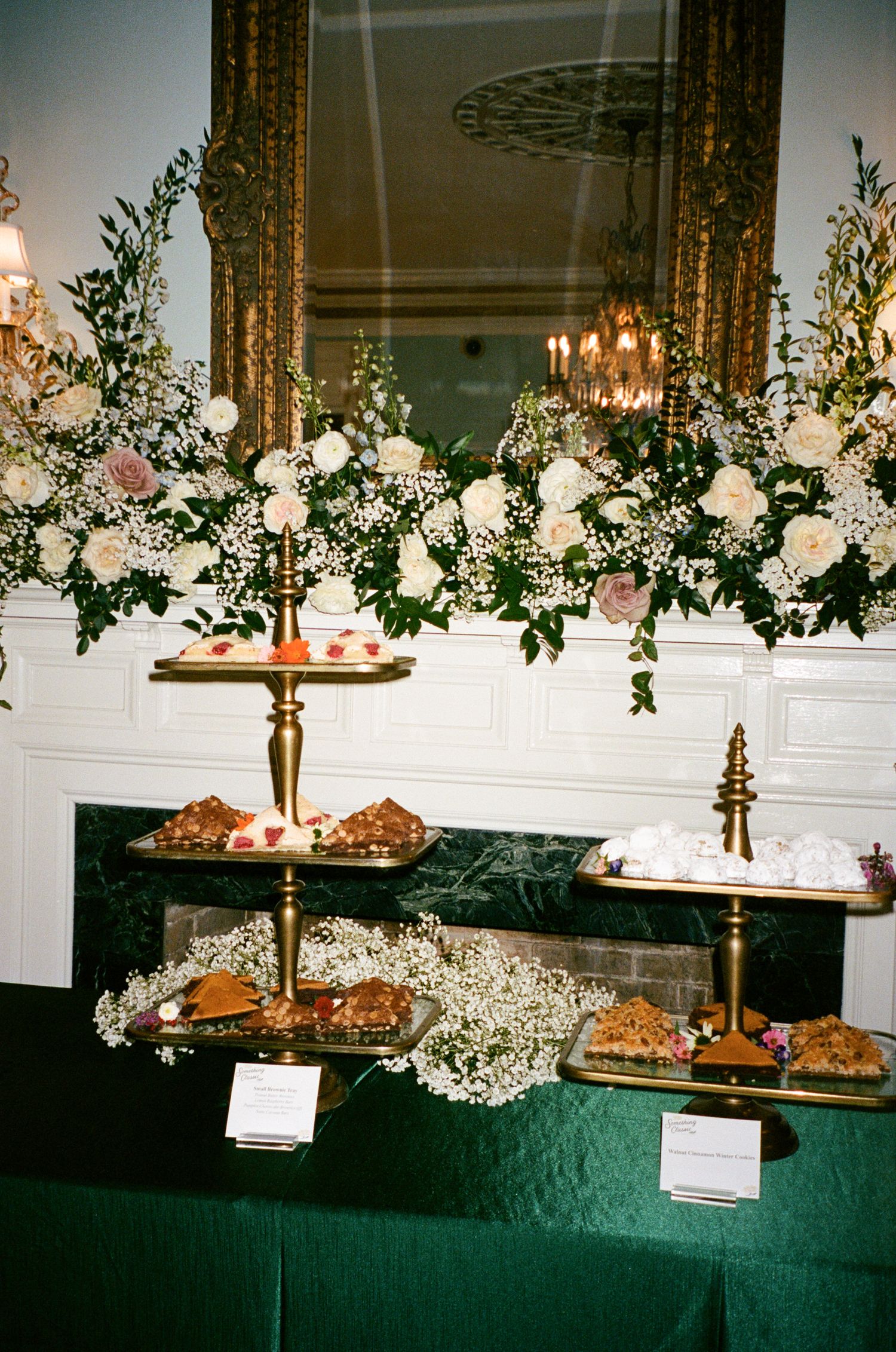 Tiered gold pastry stands with croissants and desserts on a green tablecloth beneath a white floral arrangement on an ornate mantel.