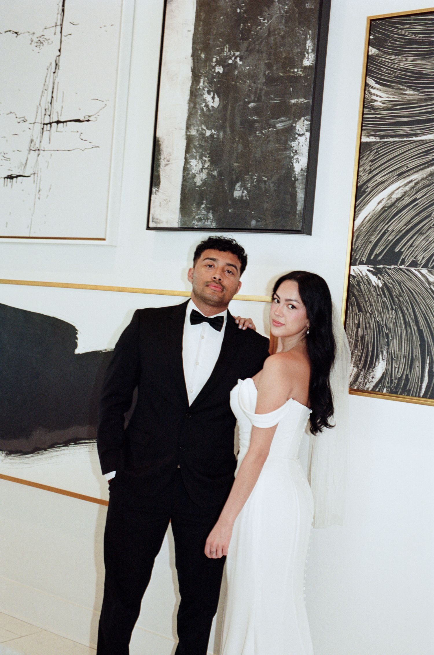 A couple poses together in front of black and white abstract artwork displayed on a white gallery wall.