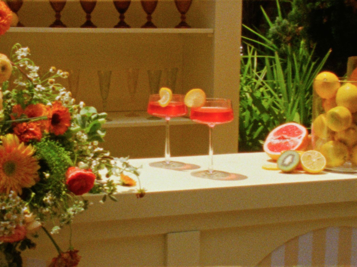 Two red cocktails garnished with lemon slices on a white bar, surrounded by fresh citrus fruits and colorful flowers.