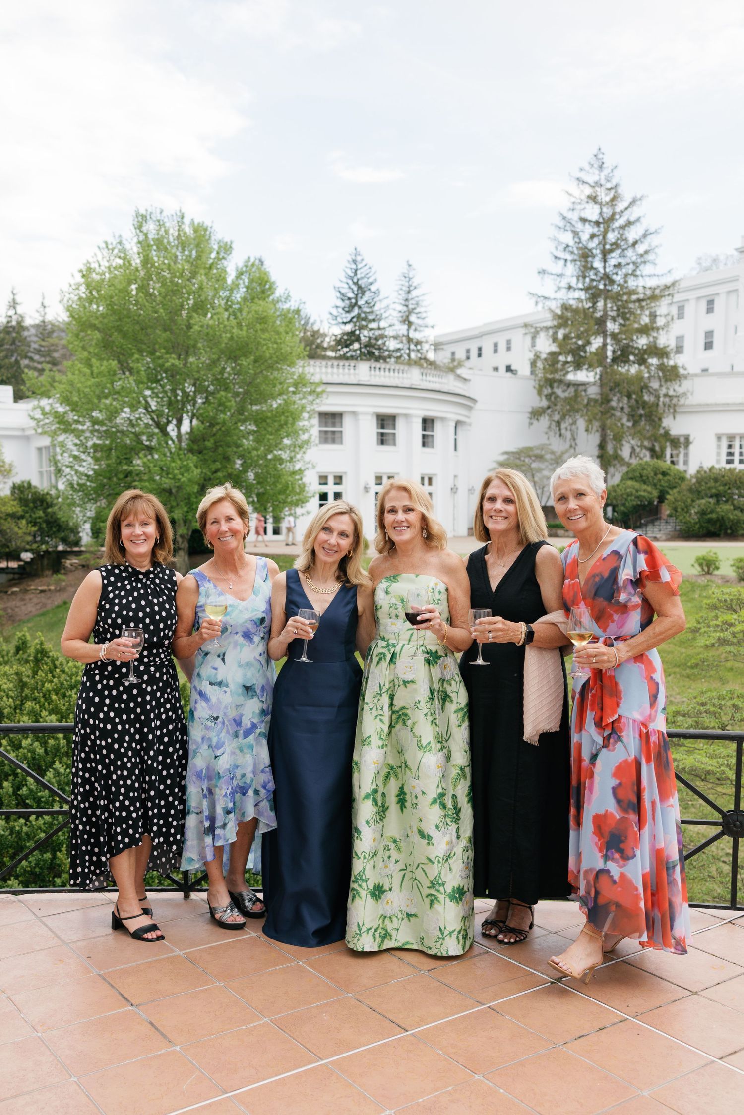 Six elegantly dressed women posing together outdoors at a garden party, holding wine glasses near a white mansion.